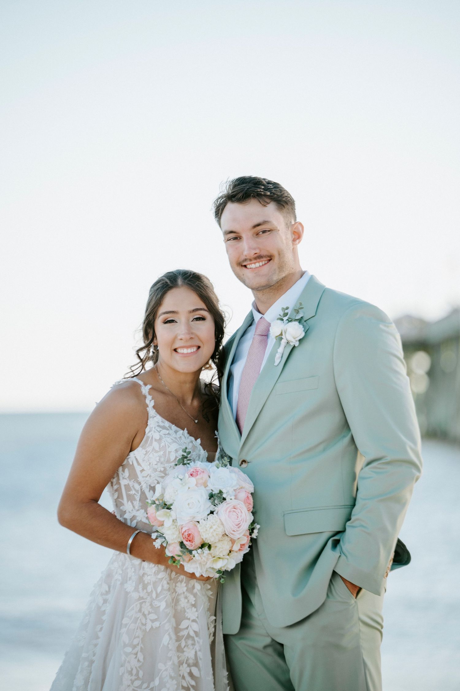 A happy couple shares tender moments on their wedding day wearing a mint green suit and lace gown by the beach.
