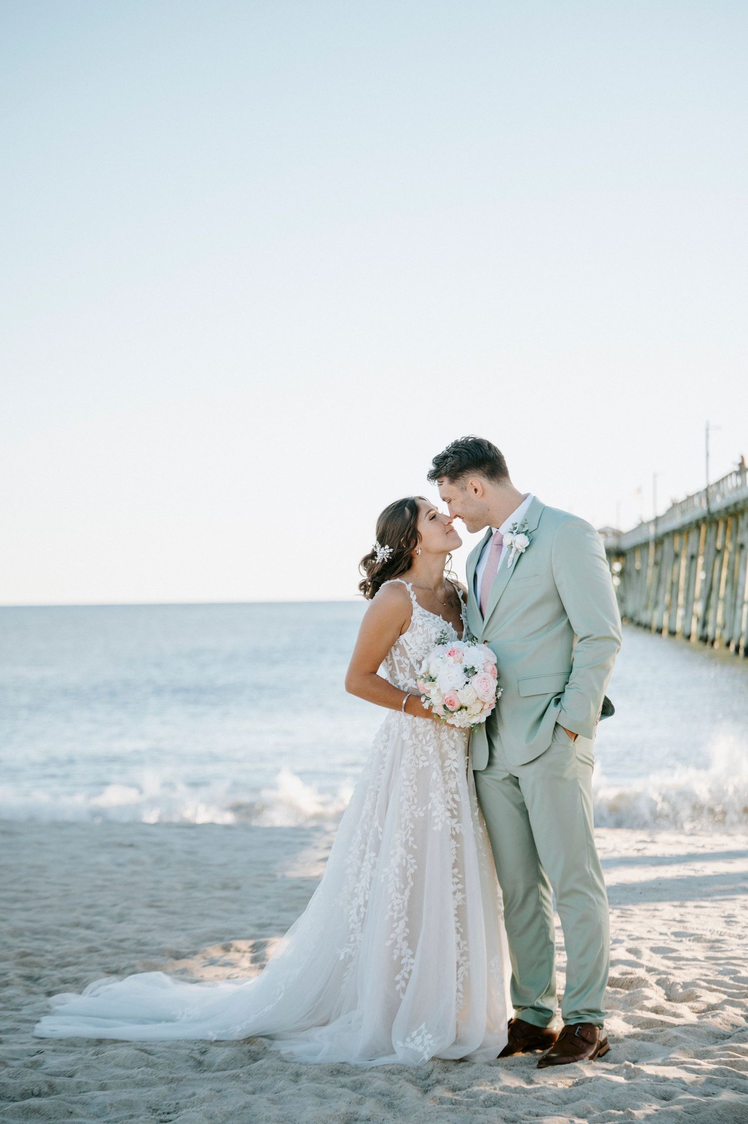 A couple shares an intimate moment together on a sandy beach with a pier and ocean waves in the background.