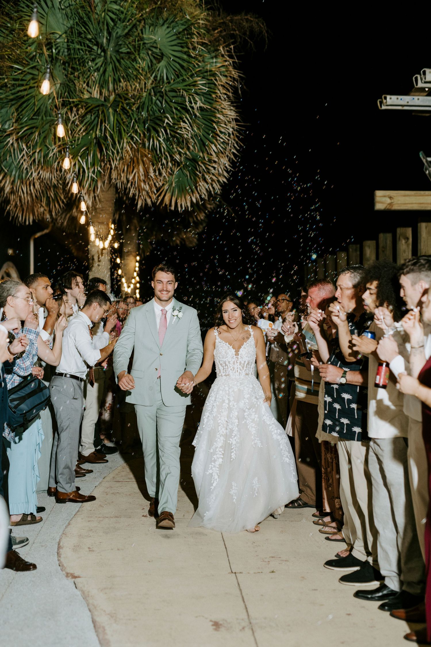 Wedding guests line up with sparklers during an evening celebration under palm trees.