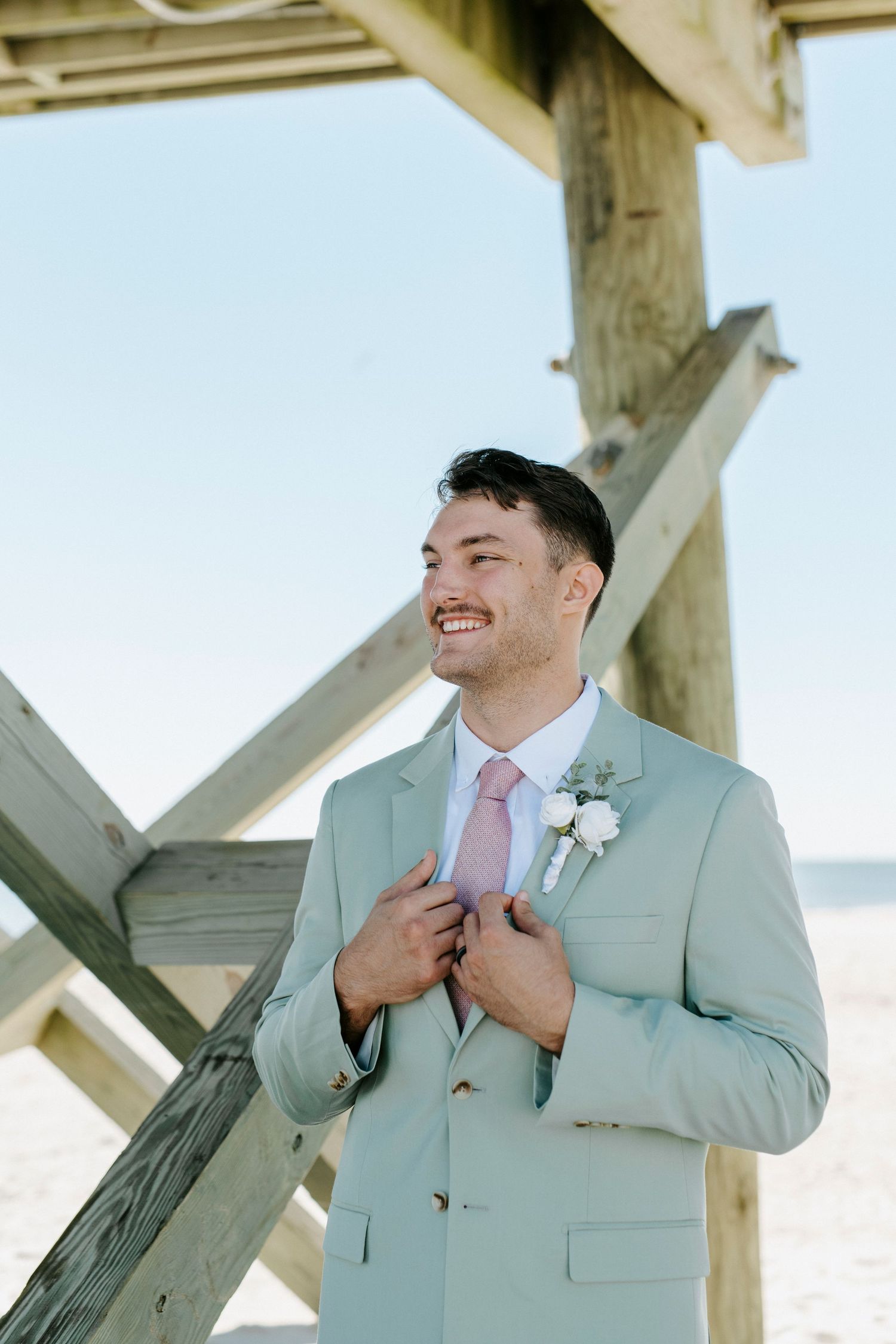 Person in light mint suit and pink tie smiles joyfully while adjusting jacket outdoors under wooden beams.