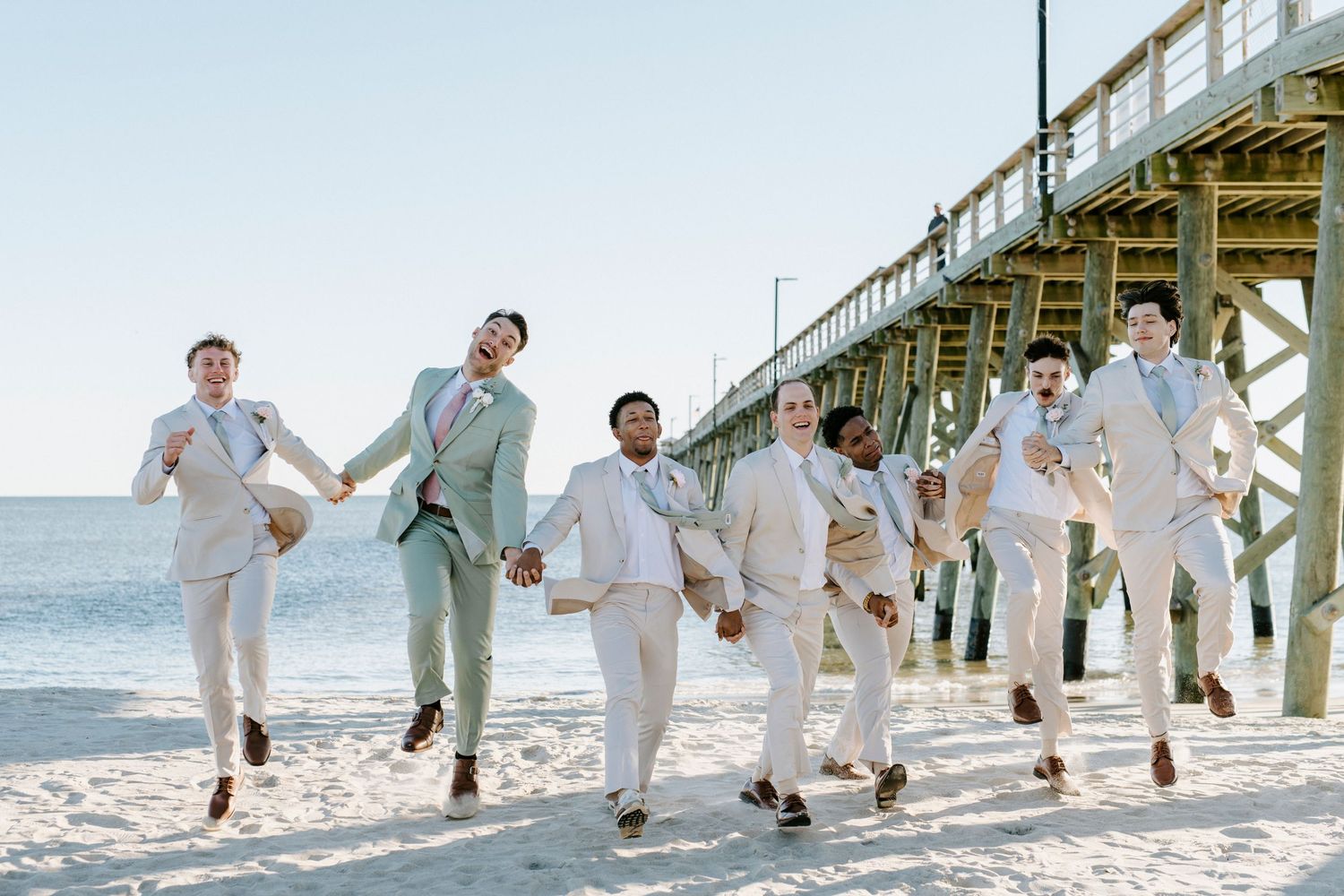 Wedding party members running and jumping on the beach in light-colored attire near a pier.