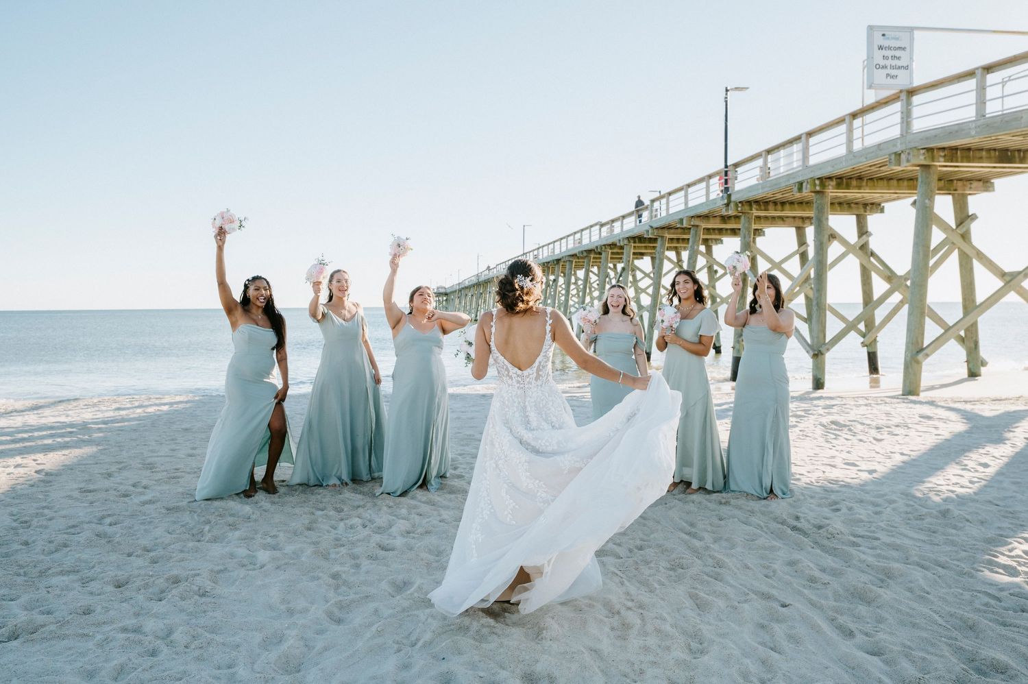 Wedding party in light blue dresses poses joyfully on a sunny beach near a wooden pier.