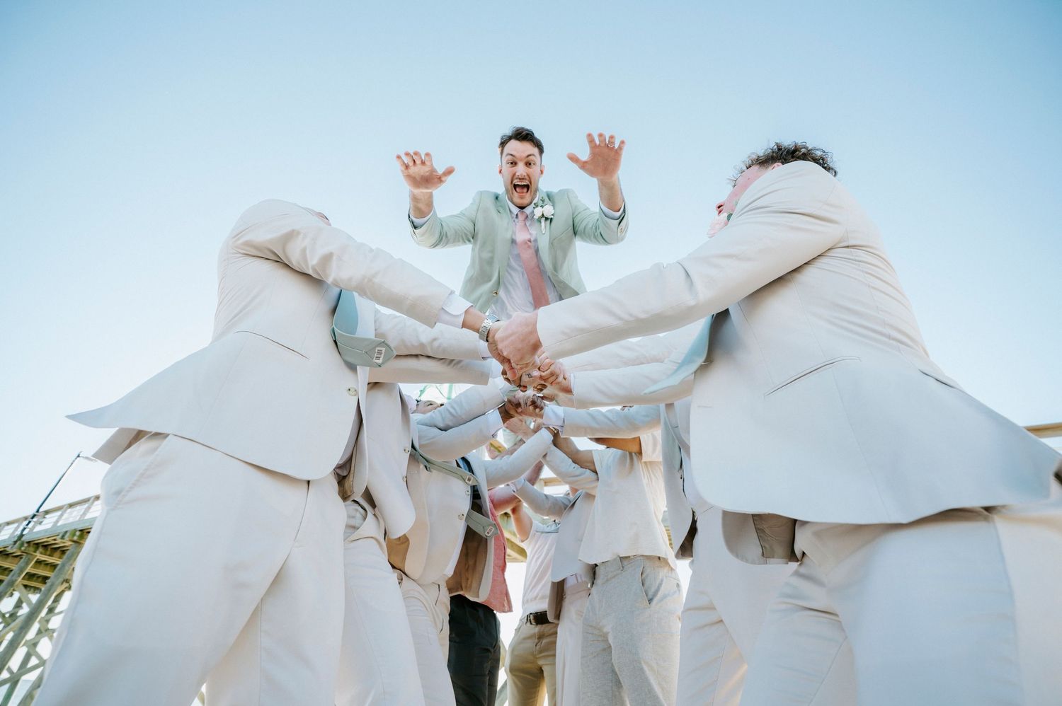 Groomsmen in light beige suits playfully toss someone in the air against a bright blue sky during a wedding celebration.