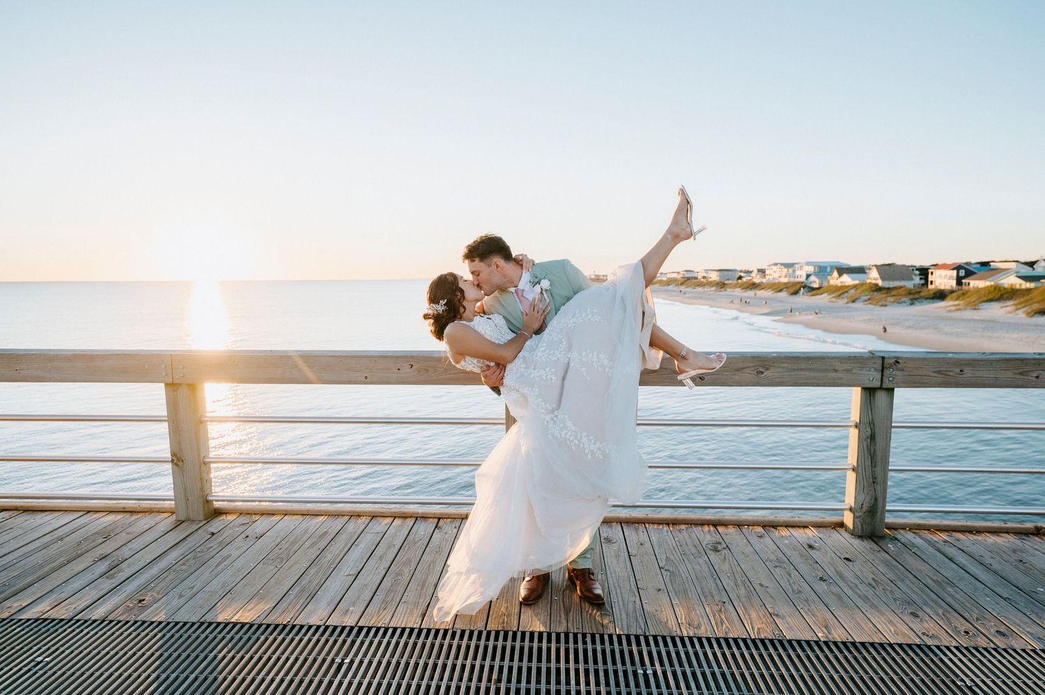 Couple in white outfits dancing romantically on a wooden pier at sunset by the ocean.