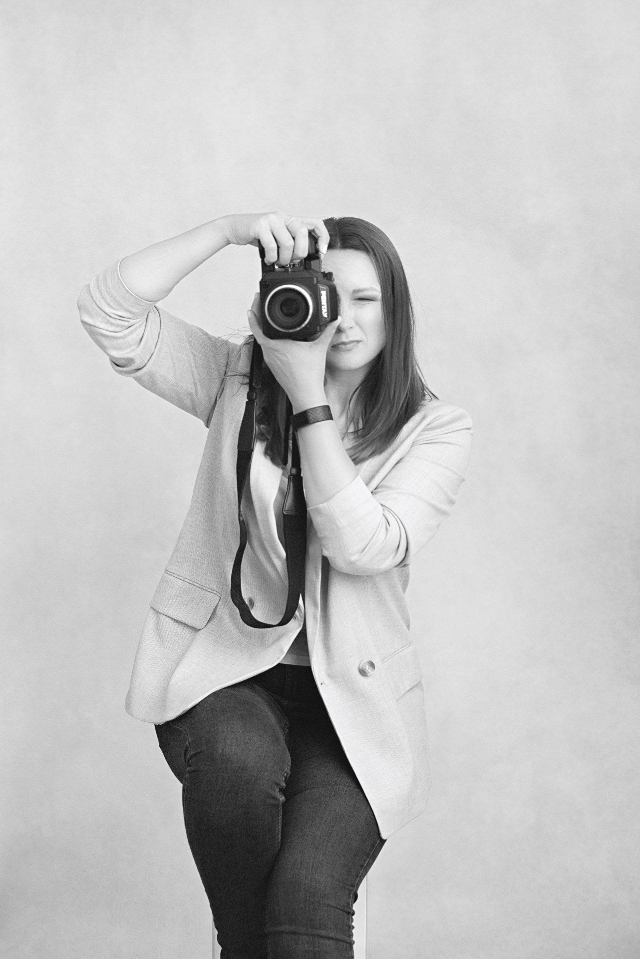 Black and white portrait of photographer taking a picture with a SLR camera while wearing a light blazer and jeans.