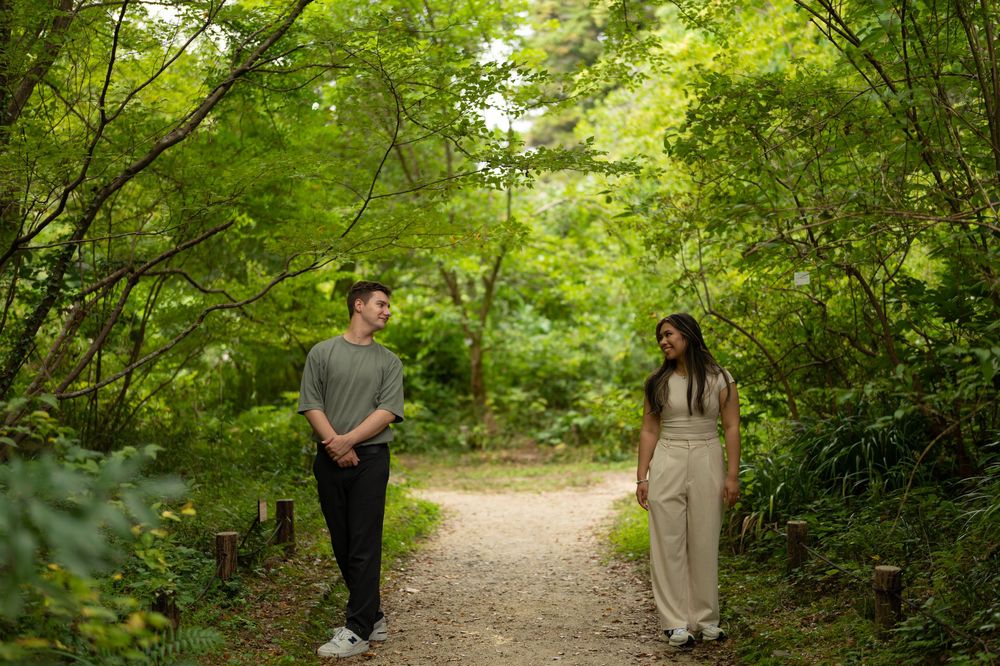 Couple smiling at each other on a winding forest path surrounded by vibrant green foliage.