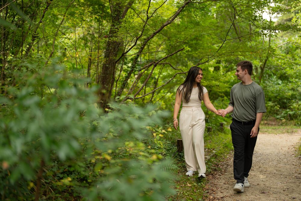 Couple holding hands walking on a forest trail surrounded by lush green trees.