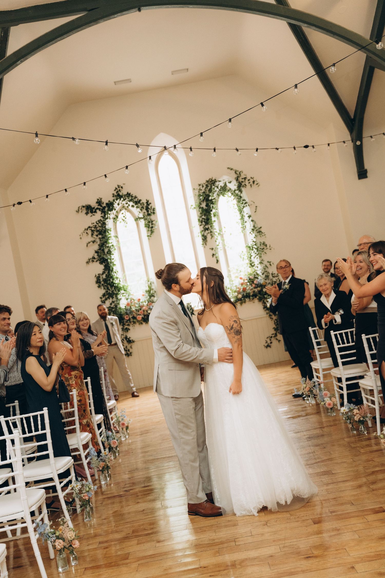 Wedding guests stand and applaud during a ceremony in a bright indoor venue decorated with string lights and greenery.