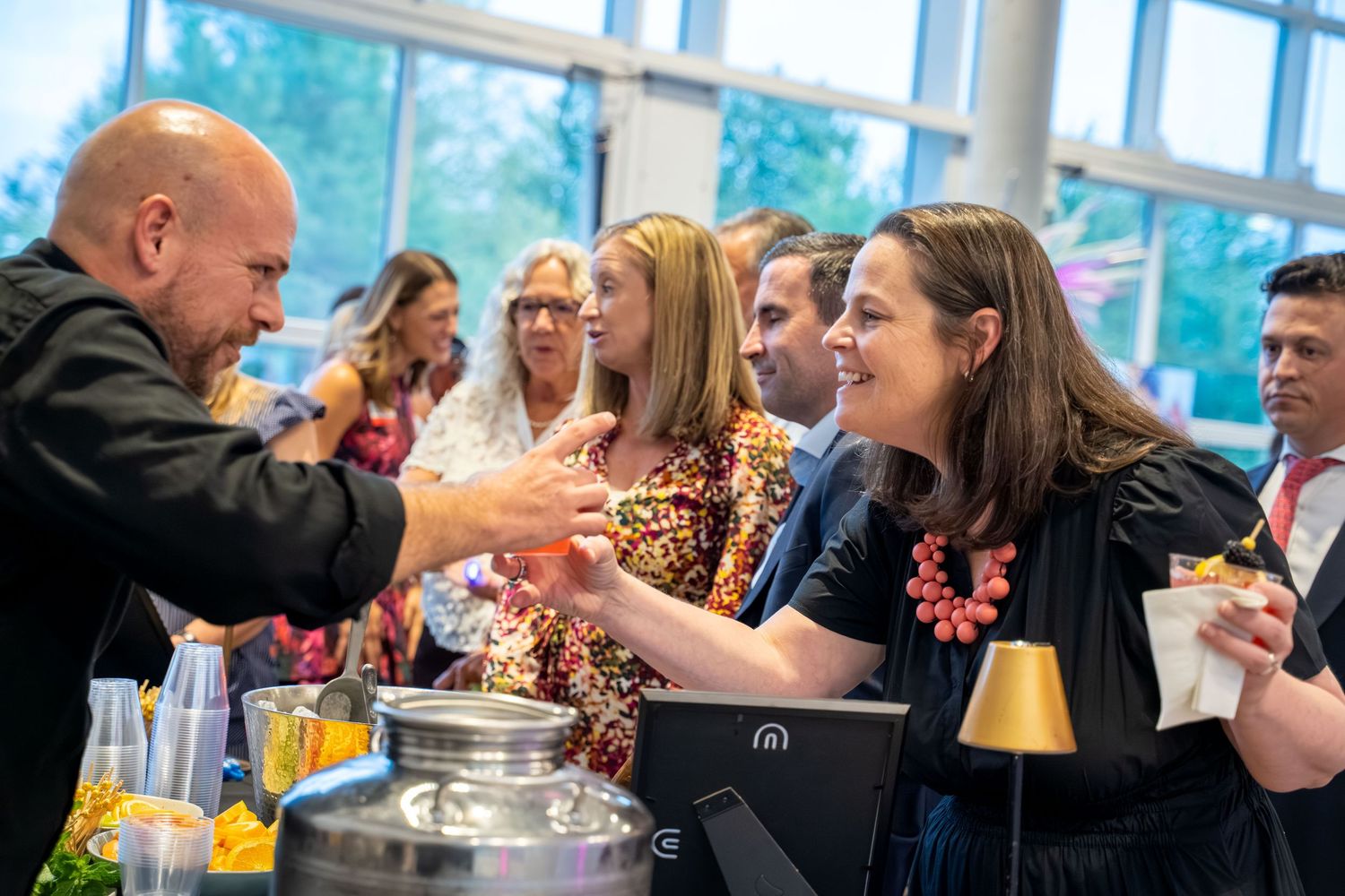 A group of people socialize and interact at a casual gathering with drinks and refreshments on the table.