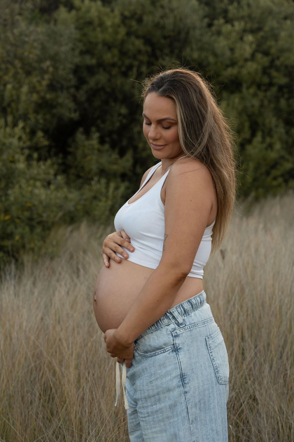 Glowing pregnant woman in denim jeans standing in tall golden grass – Wollongong maternity photography