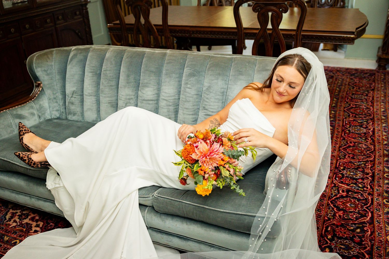 A bride in a white wedding dress lounges elegantly on a blue velvet sofa with colorful flowers nearby.