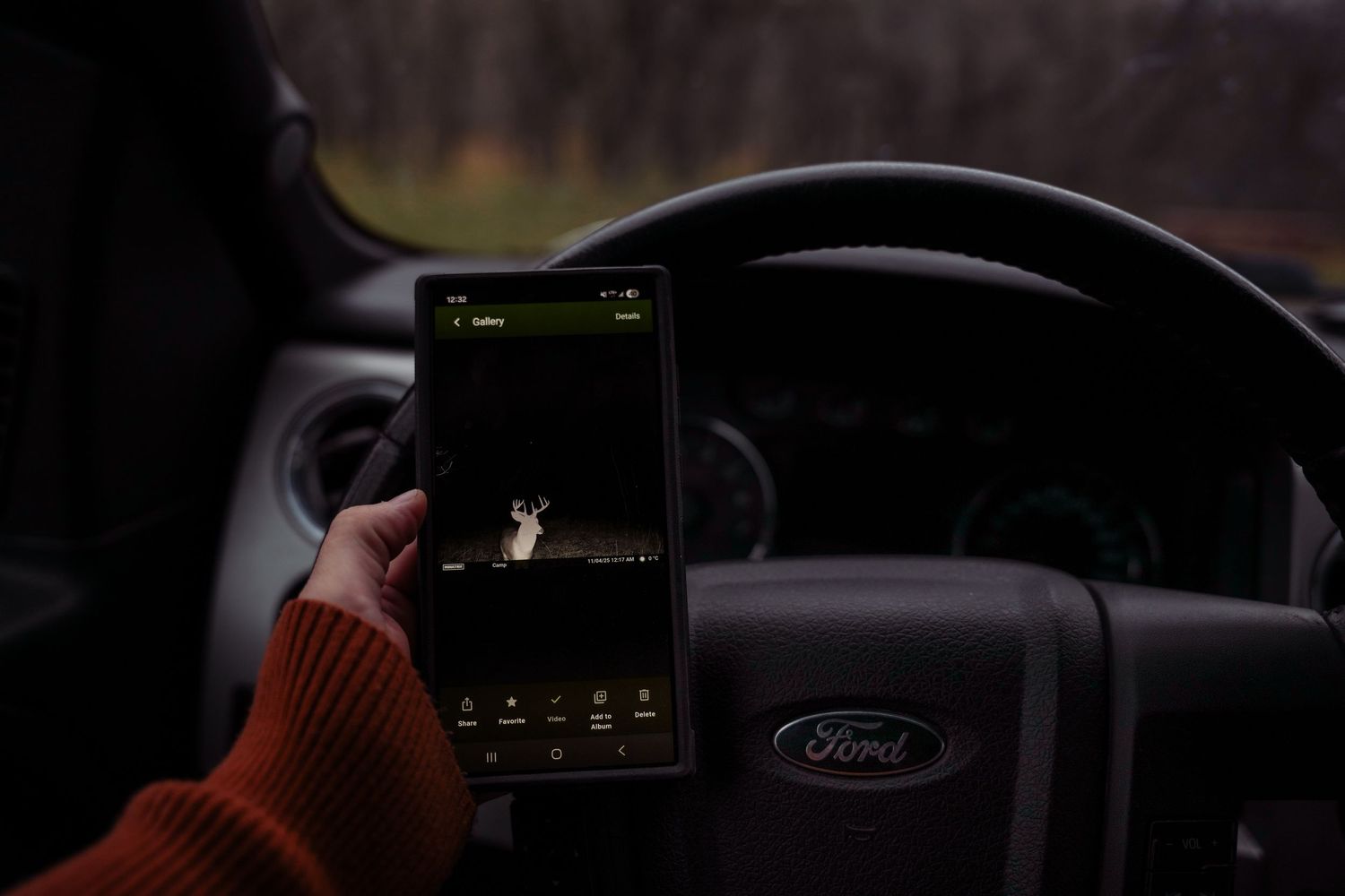 Person in orange sweater holds smartphone displaying dark screen while sitting in Ford vehicle interior.