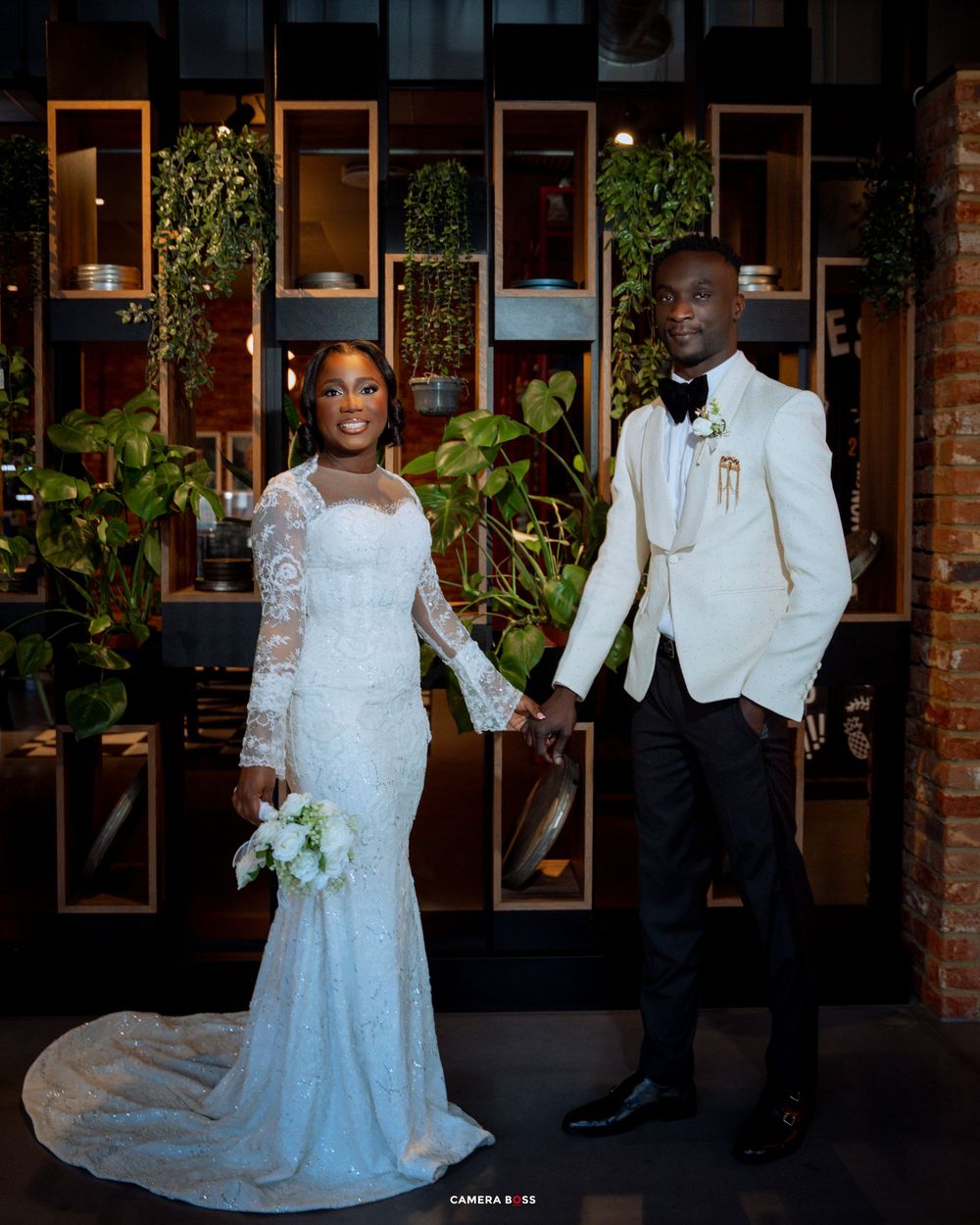 A wedding couple poses together in a rustic brick-walled venue with greenery and warm lighting in the background.
