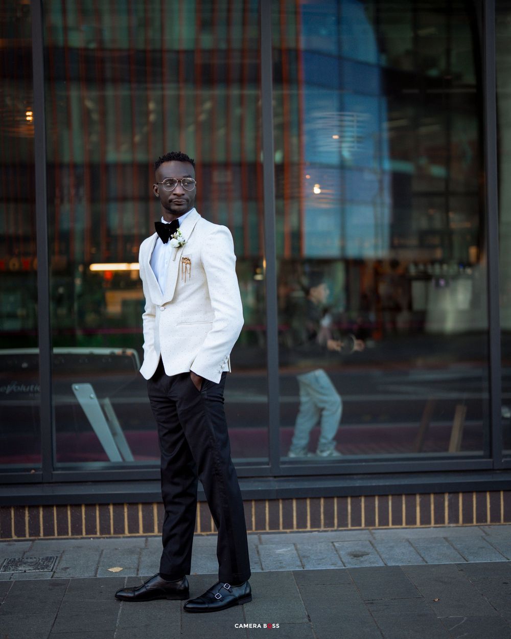Person in crisp white tuxedo jacket and black pants poses outdoors near modern building windows.