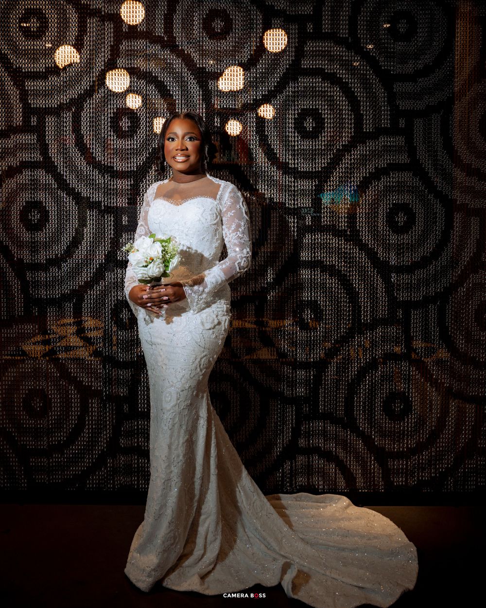 Elegant bride in a long-sleeved lace wedding gown poses against a dark patterned wall.