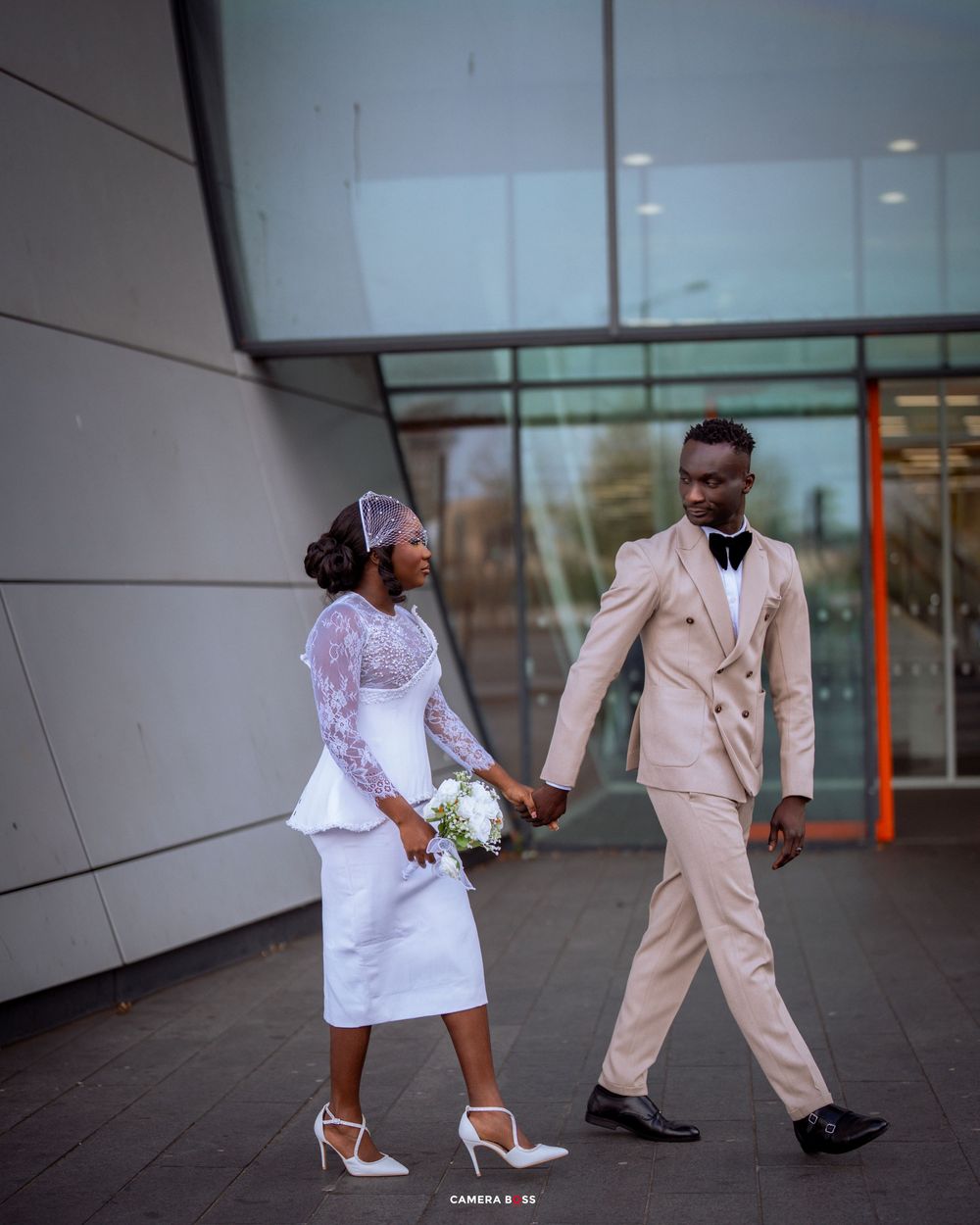 Stylish couple in white and beige outfits poses together outside a modern building during a wedding photoshoot.