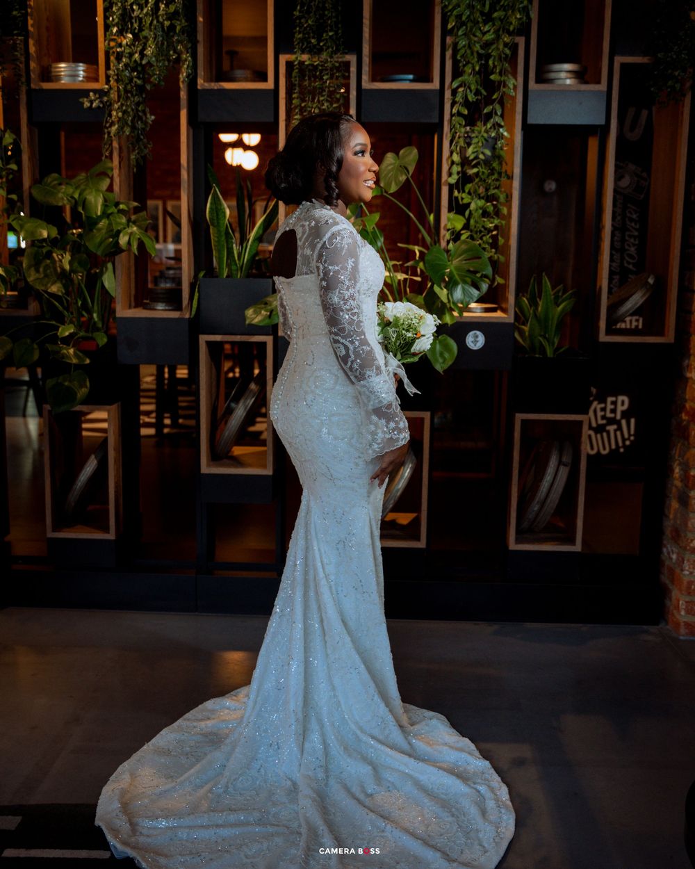A beautiful white lace wedding dress is showcased in an elegant indoor setting with dark walls and framed artwork.