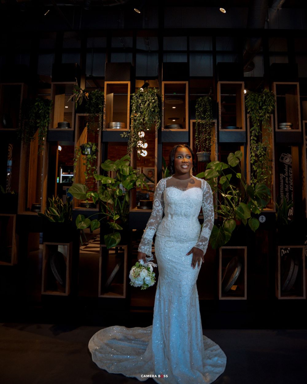 A bride in a white lace gown poses elegantly in front of a dark background with illuminated windows.