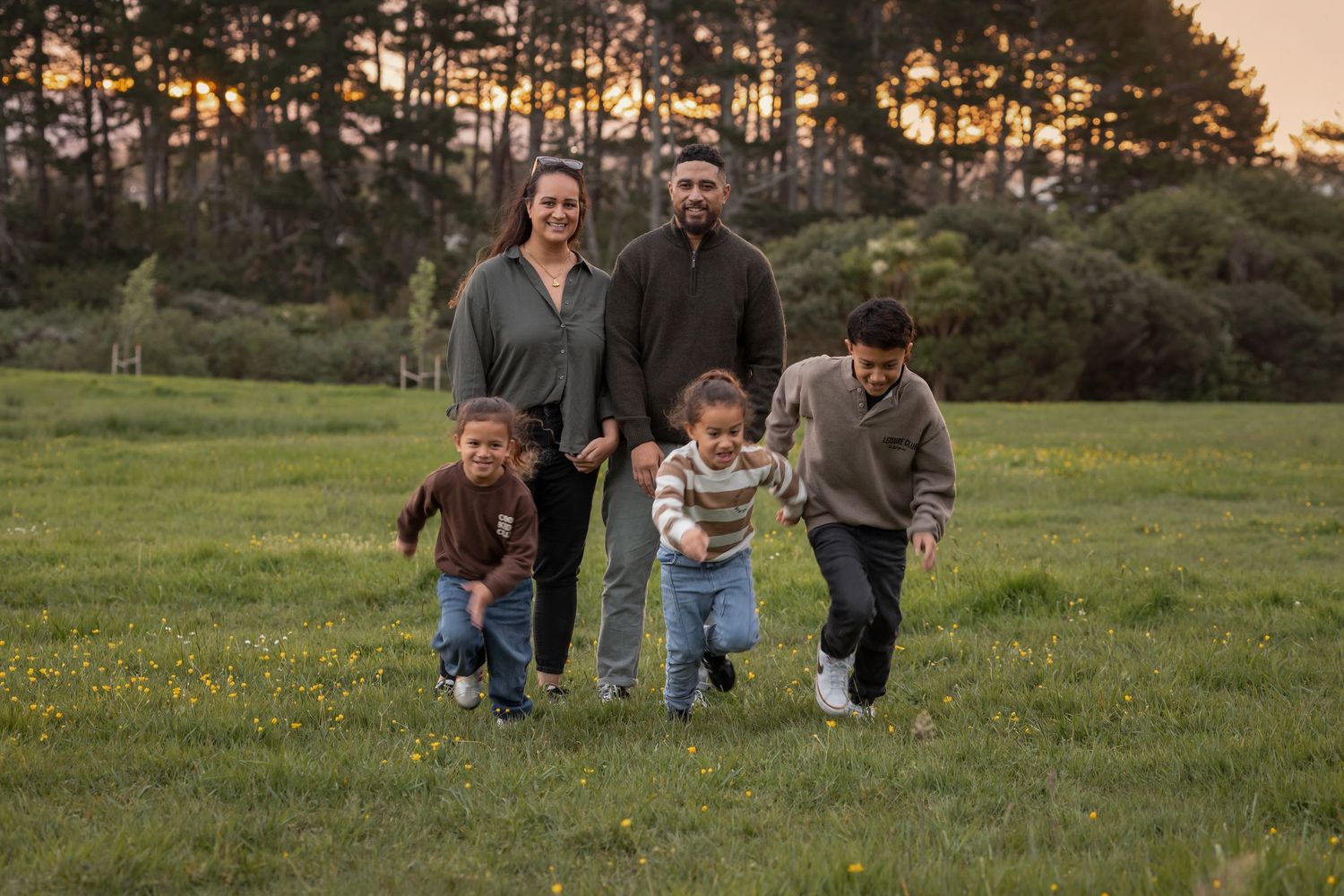 Outdoor family phot on a grass field. Parents stand behind 3 children who run towards the camera. Large trees in the background with sunset