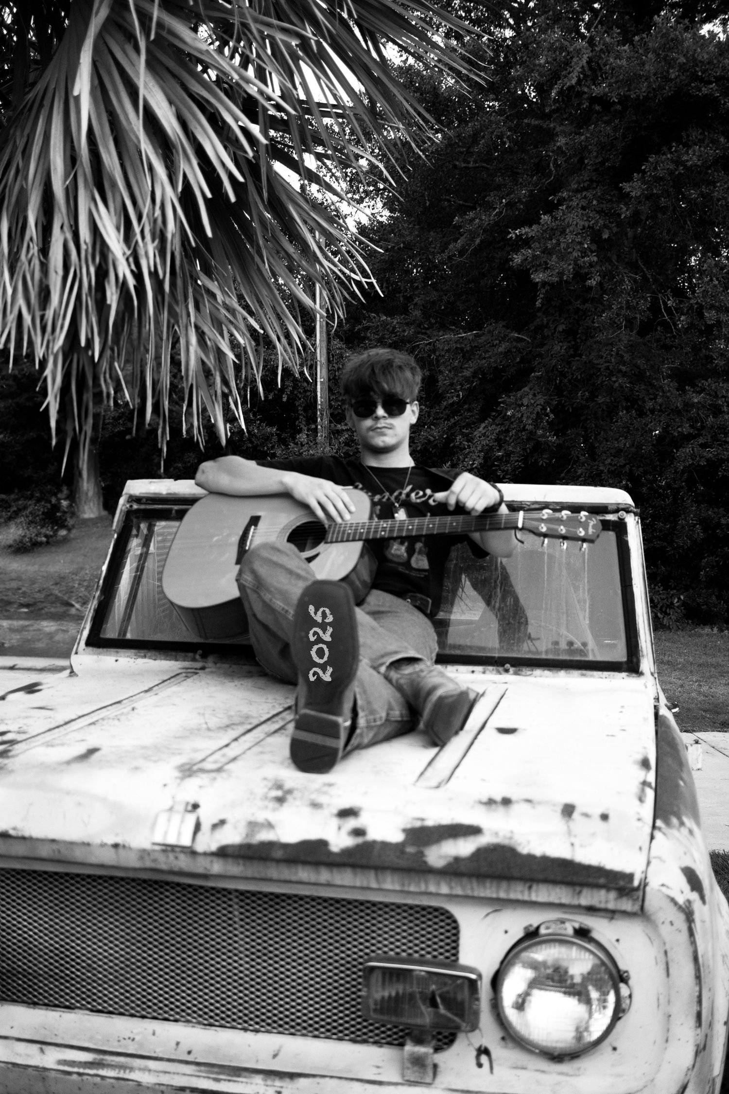 Black and white image of someone playing acoustic guitar while sitting on an old truck hood under palm trees.