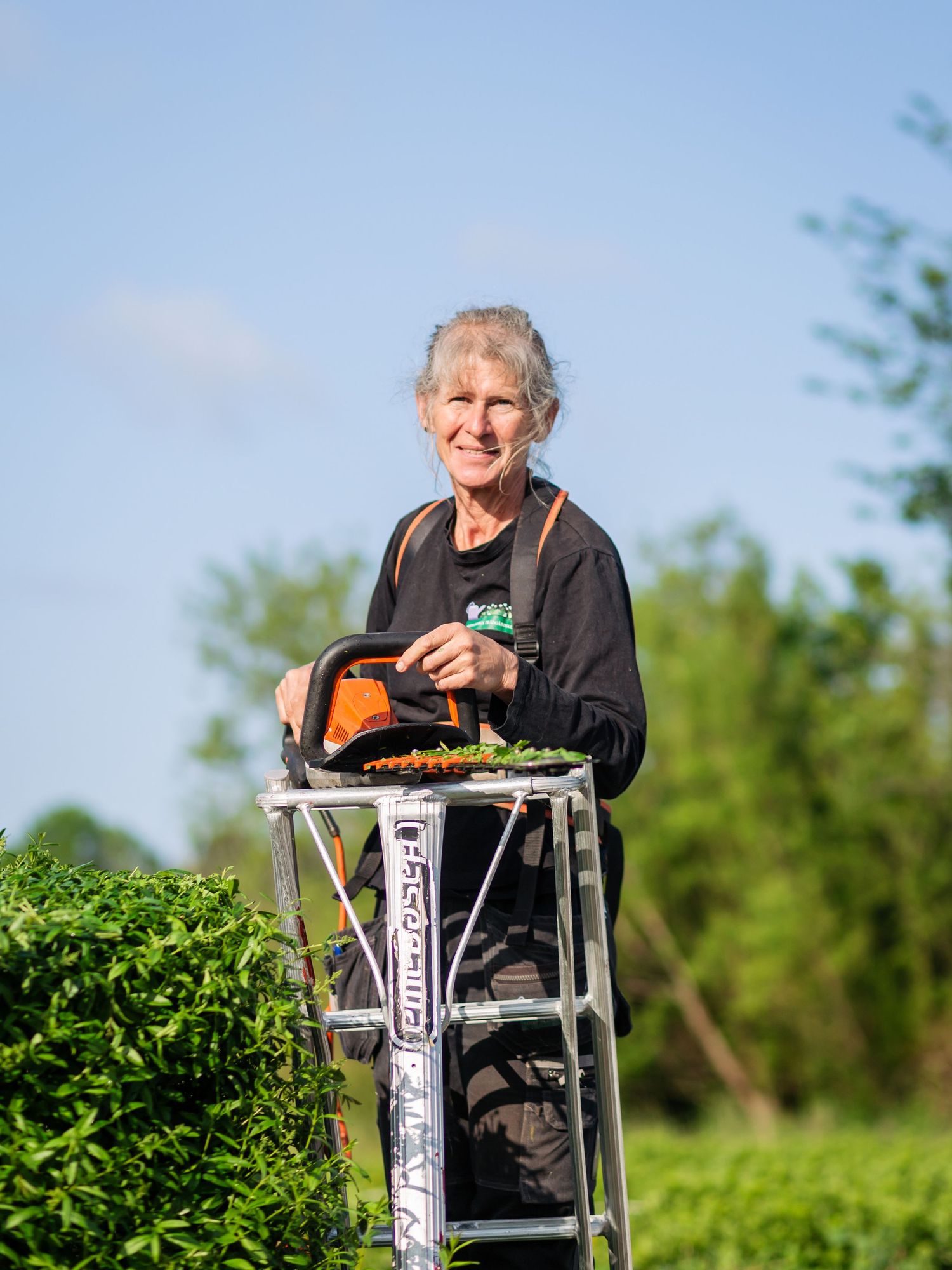 Person in black clothing sits in a tall chair outdoors enjoying a meal against a backdrop of green foliage.