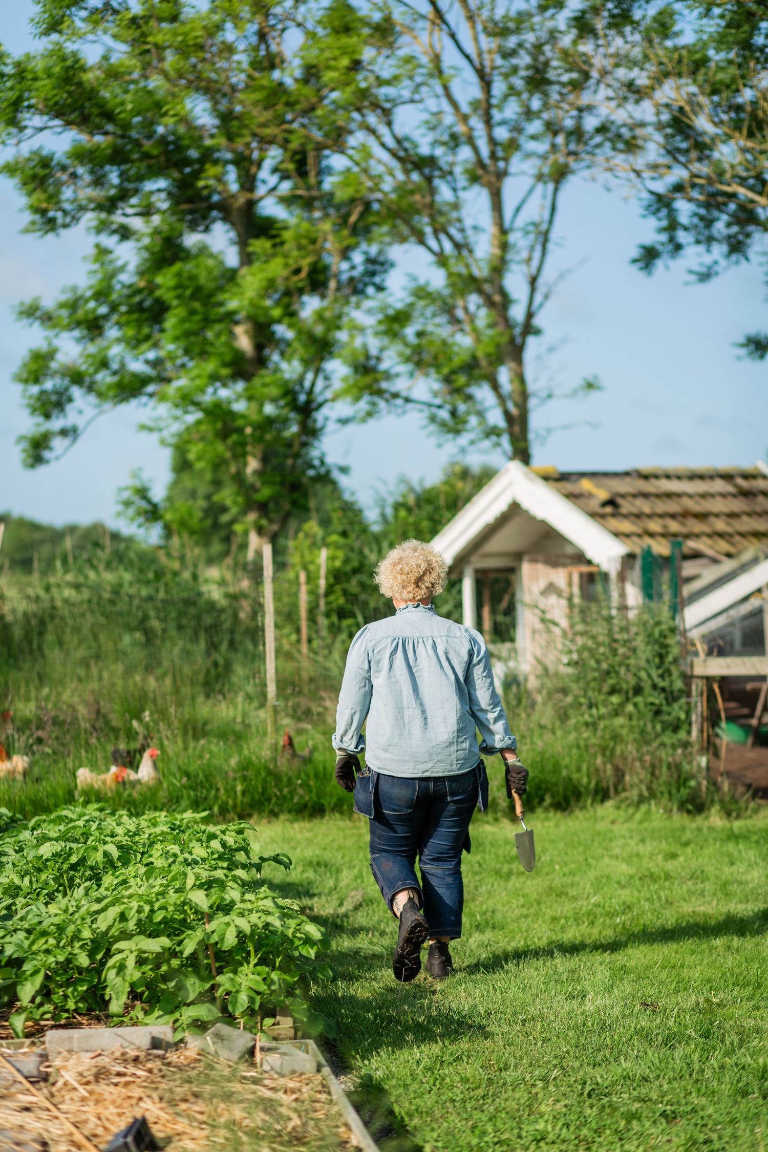 A person in denim overalls walks through a lush green garden with trees and small structures in the background.