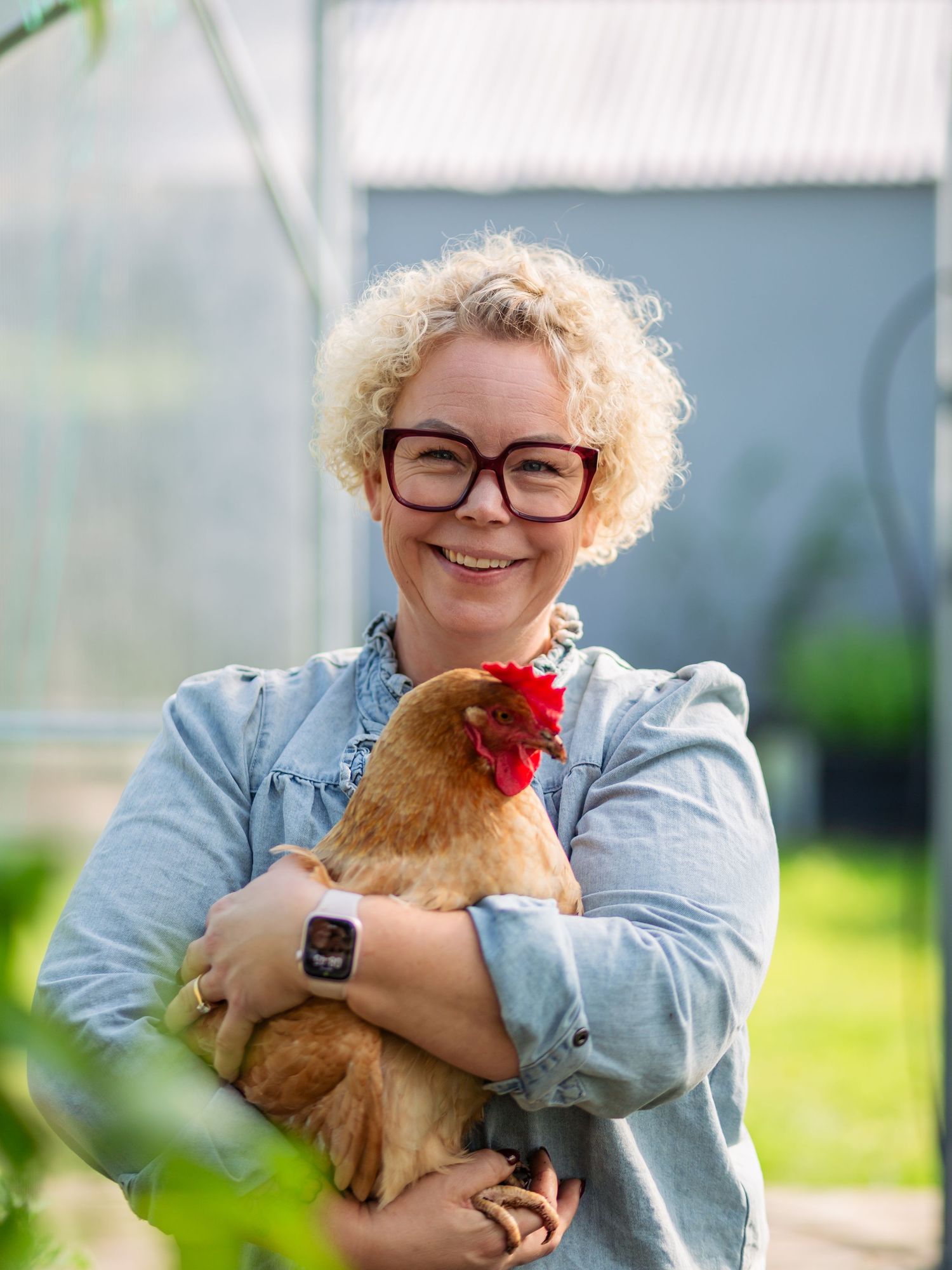 Someone wearing glasses and a denim jacket holds a brown chicken with red comb while standing outdoors.