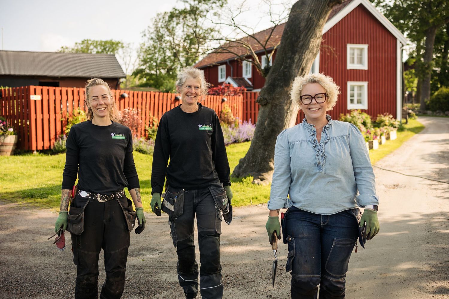 Three people walking together on a gravel path with traditional red Swedish buildings in the background.
