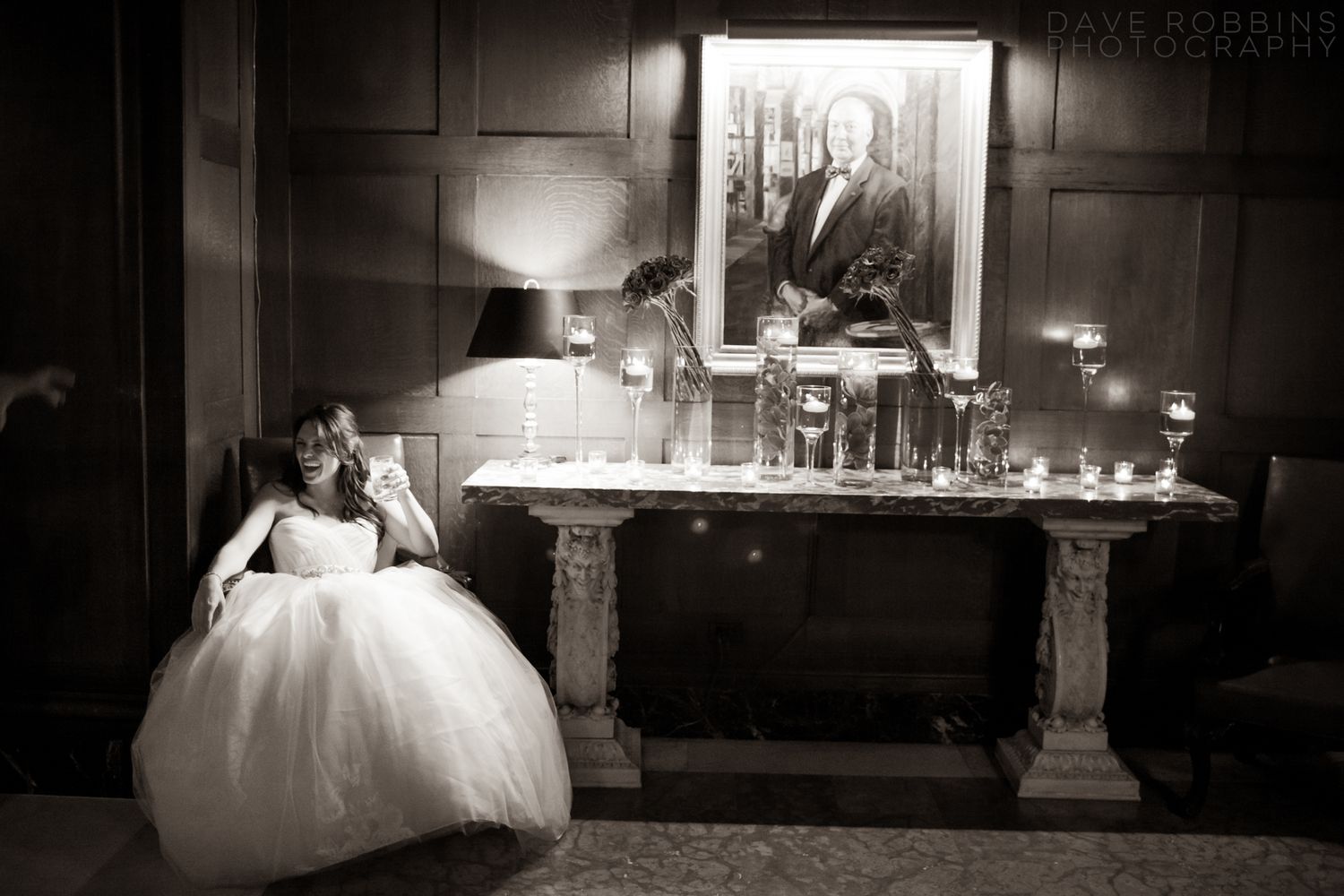 A bride in a white ball gown sits elegantly beside an ornate fireplace mantel in a dramatic black and white photograph.