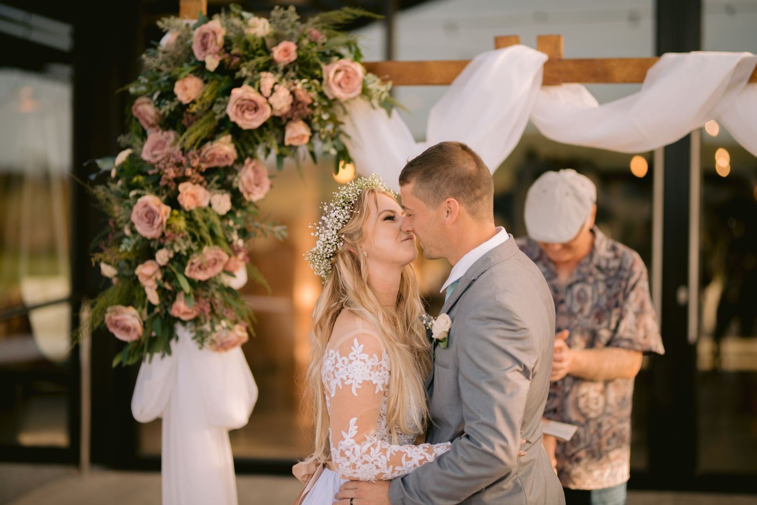 Romantic wedding couple embraces under pink floral arch with soft draping and string lights at sunset.
