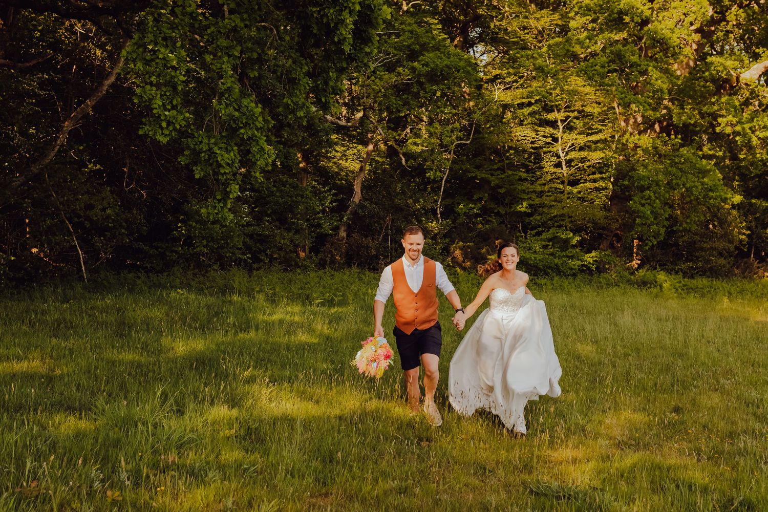 Newlyweds running hand in hand across a grassy field, joyful and playful.