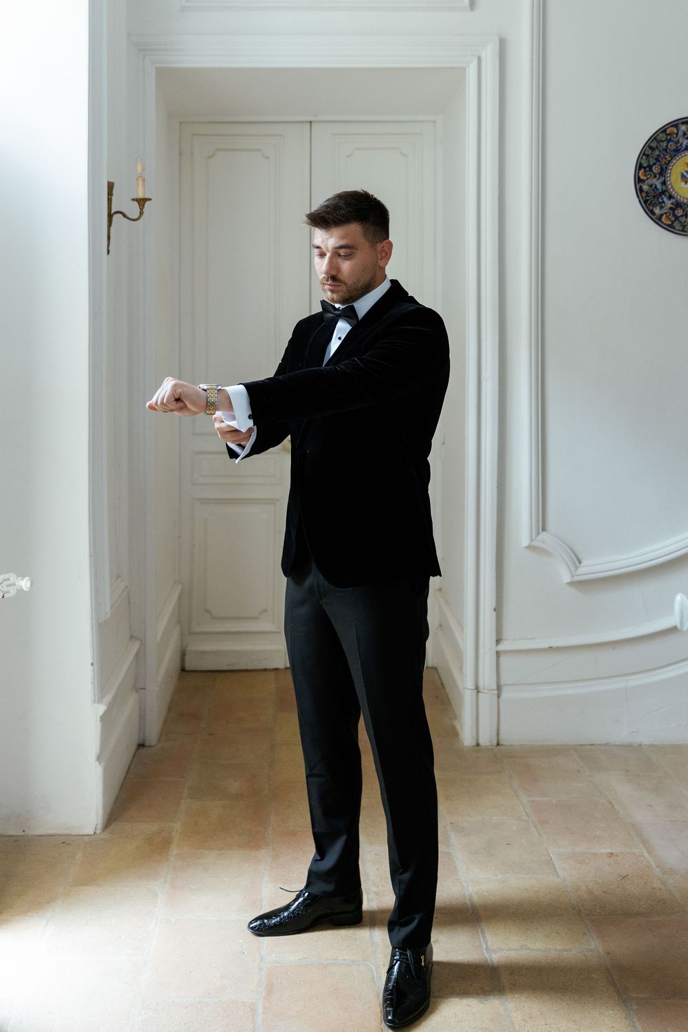 Elegant figure in black tuxedo and bow tie checking time while standing in a classic white interior hallway.