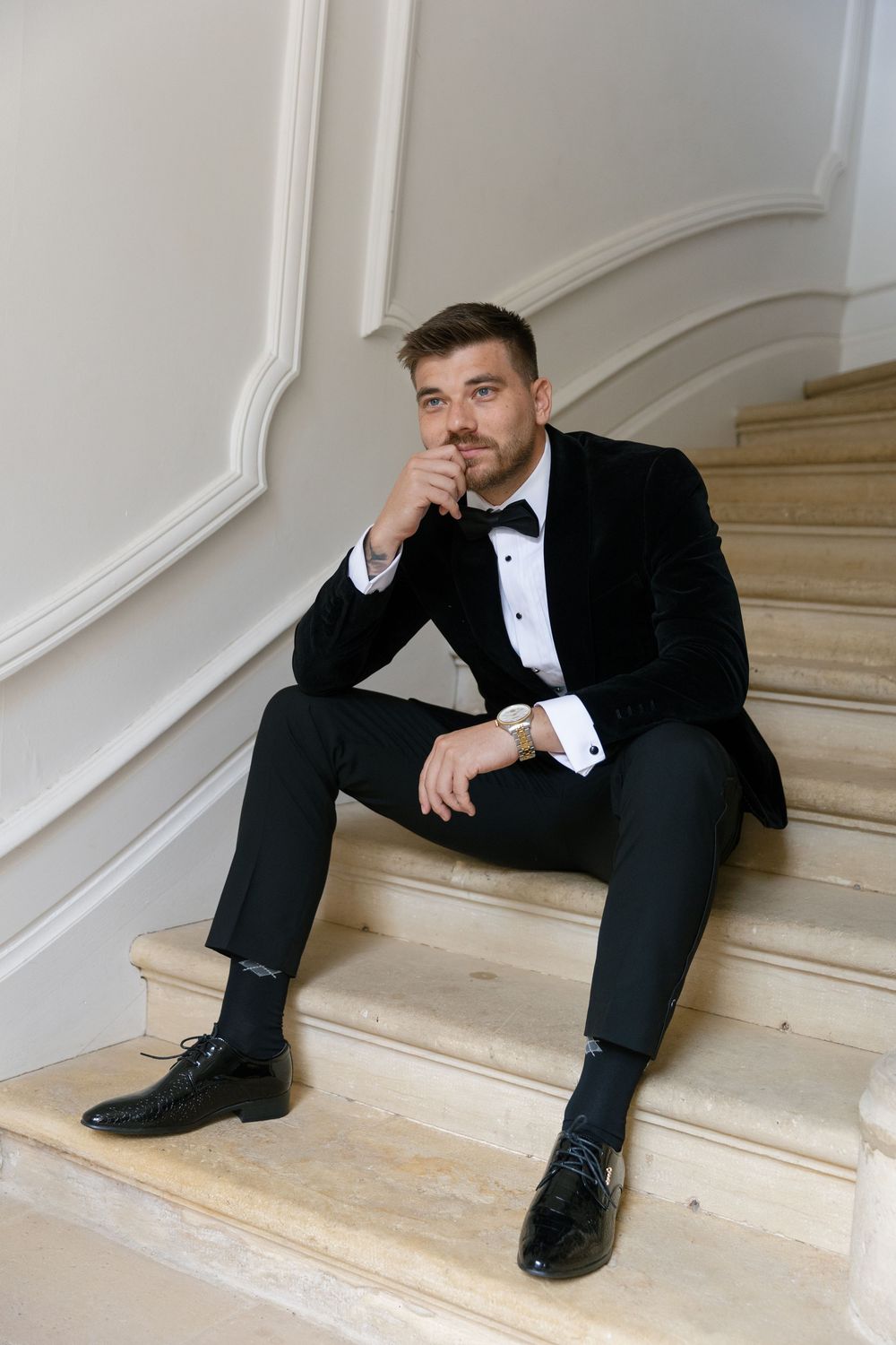 Formal figure in tuxedo sitting thoughtfully on marble stairs in an elegant white interior setting.