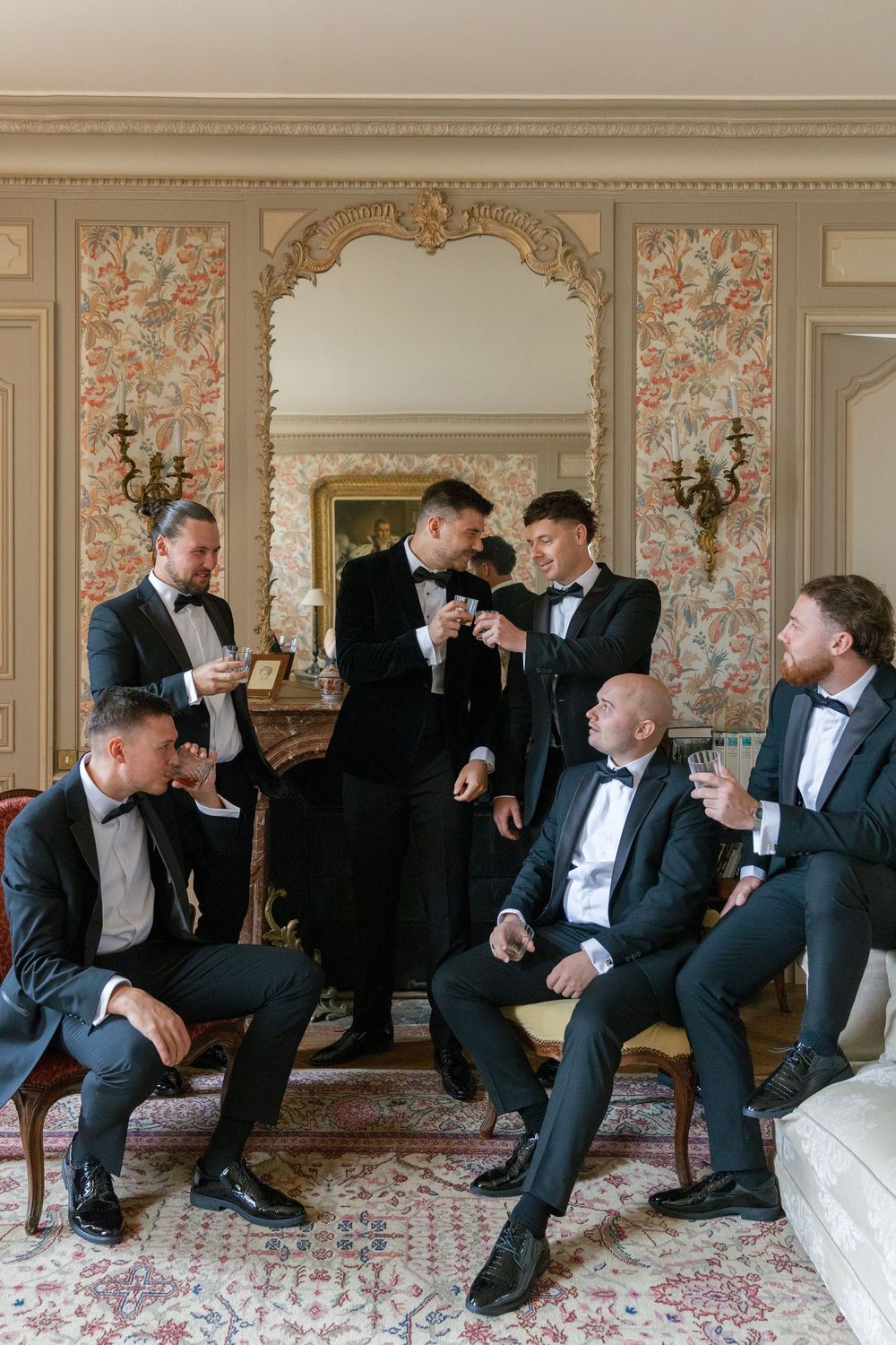 Group of groomsmen in tuxedos relaxing on ornate furniture in an elegant room with floral wallpaper.