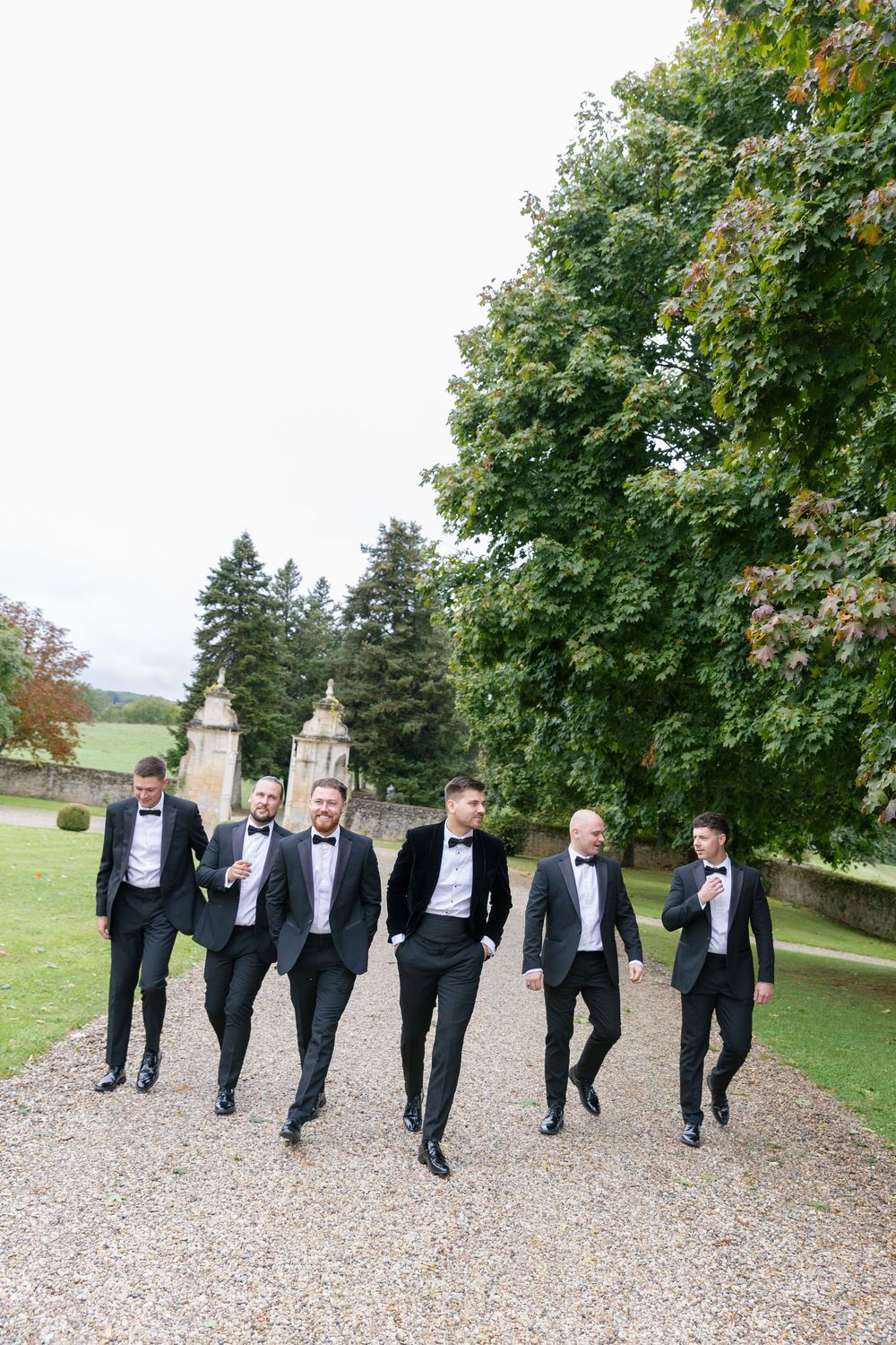 A group of groomsmen in black tuxedos walking together on a gravel path surrounded by trees.