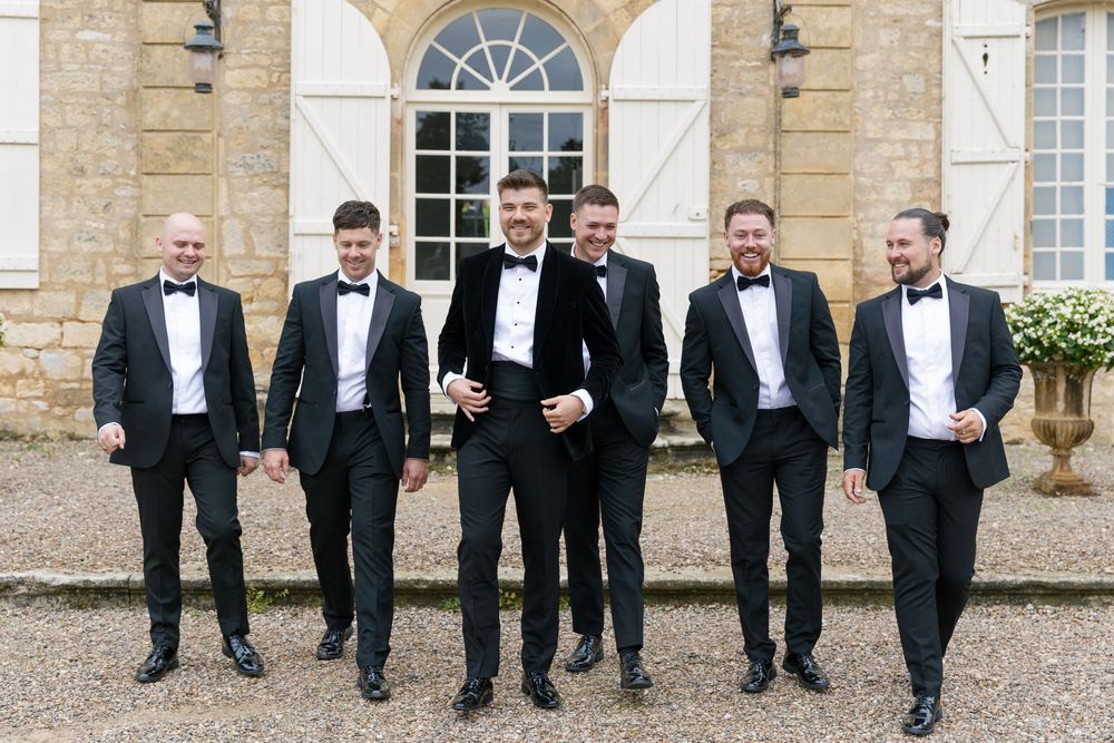 A group in black tuxedos and bow ties stand together smiling outside an elegant stone building with arched windows.