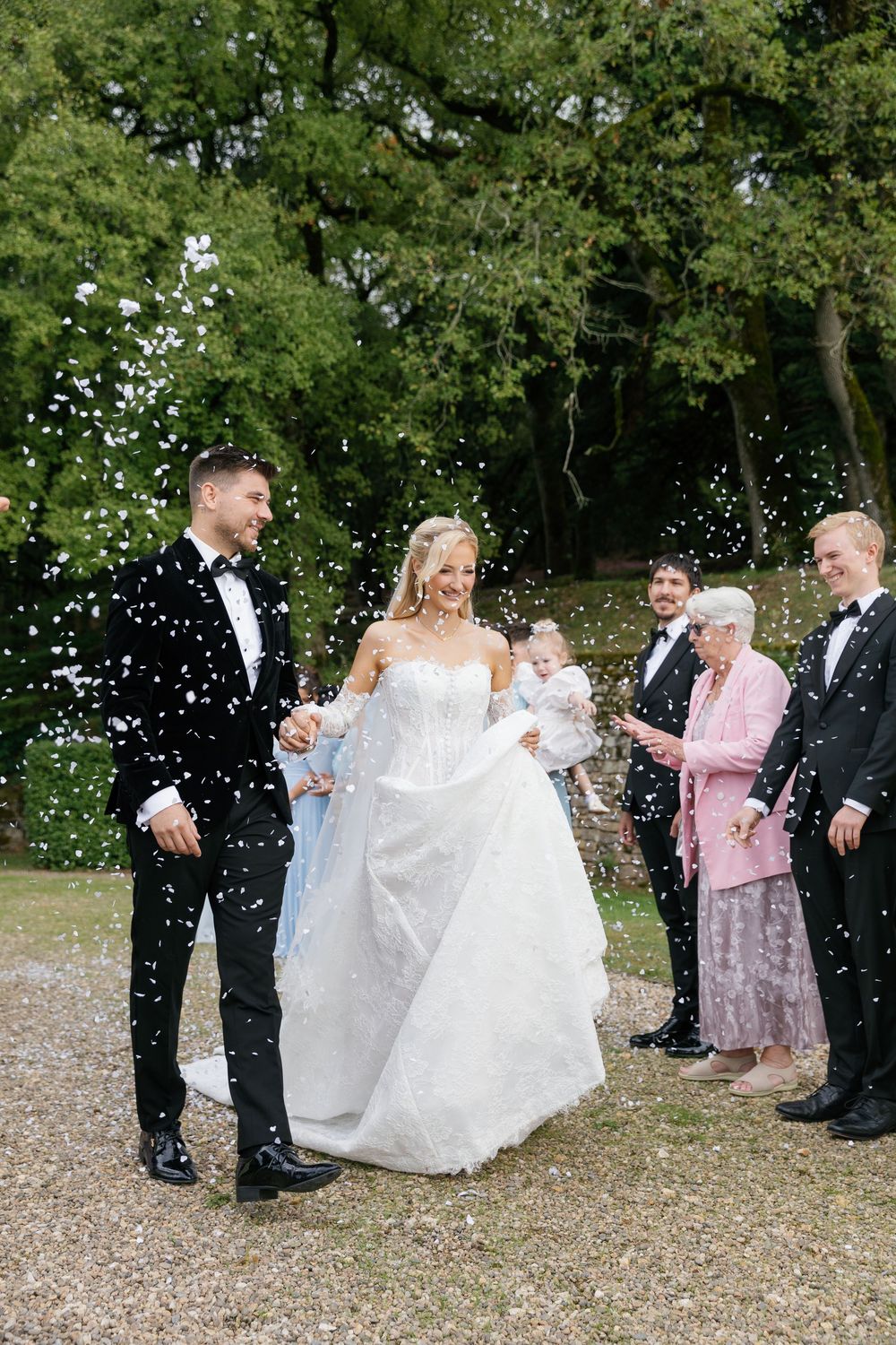 Wedding celebration on garden grounds with guests throwing confetti amid green trees.