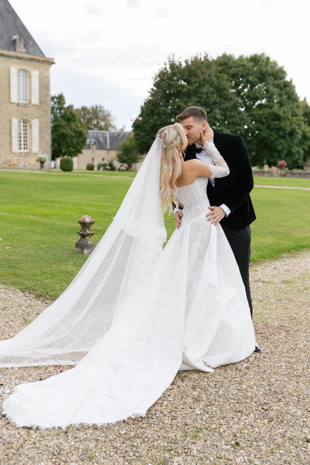 Romantic wedding portraits on gravel path with flowing white dress train against chateau backdrop.