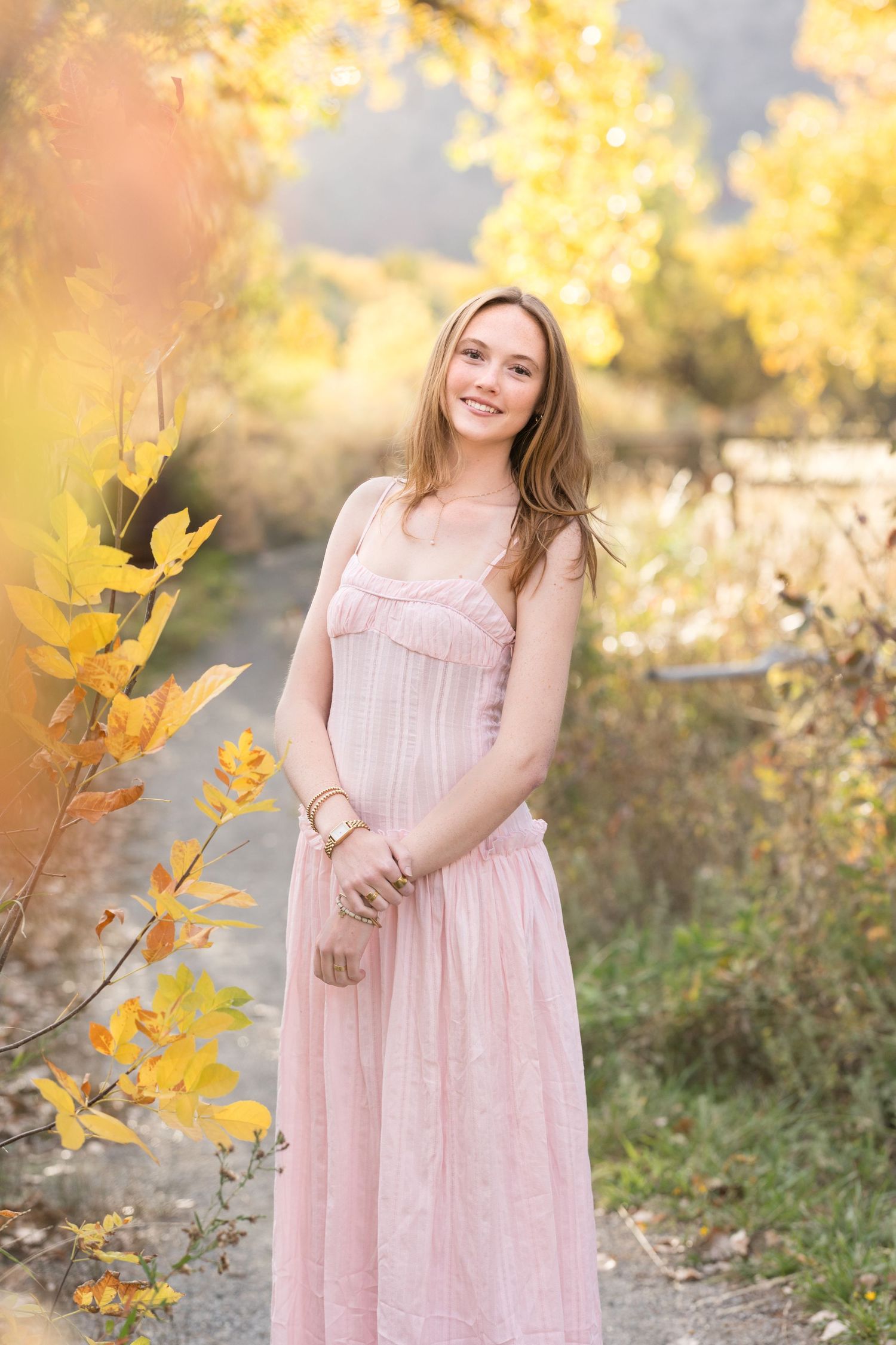 A person in a pink maxi dress poses outdoors during autumn with golden foliage in the background.