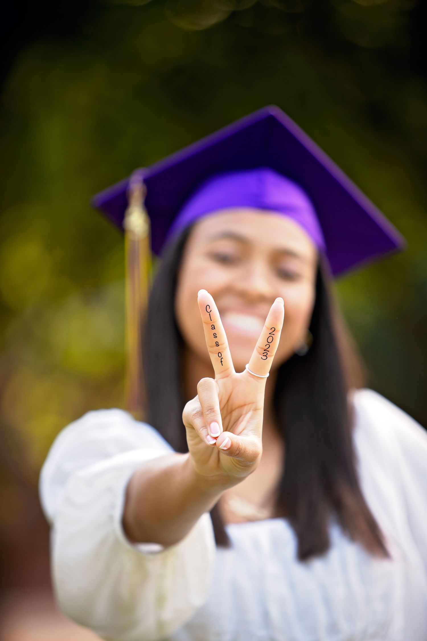 Graduate in purple cap and gown making peace sign gestures in natural outdoor setting.