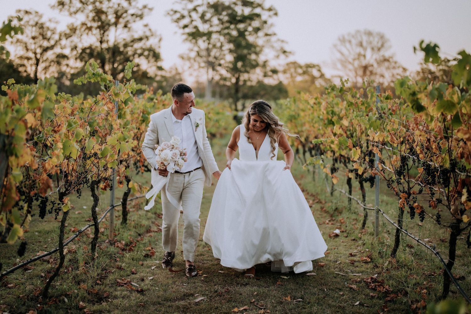 Romantic couple walking through a vineyard at sunset in wedding attire with flowing white dress and light colored suit at Enzo Hunter Valley