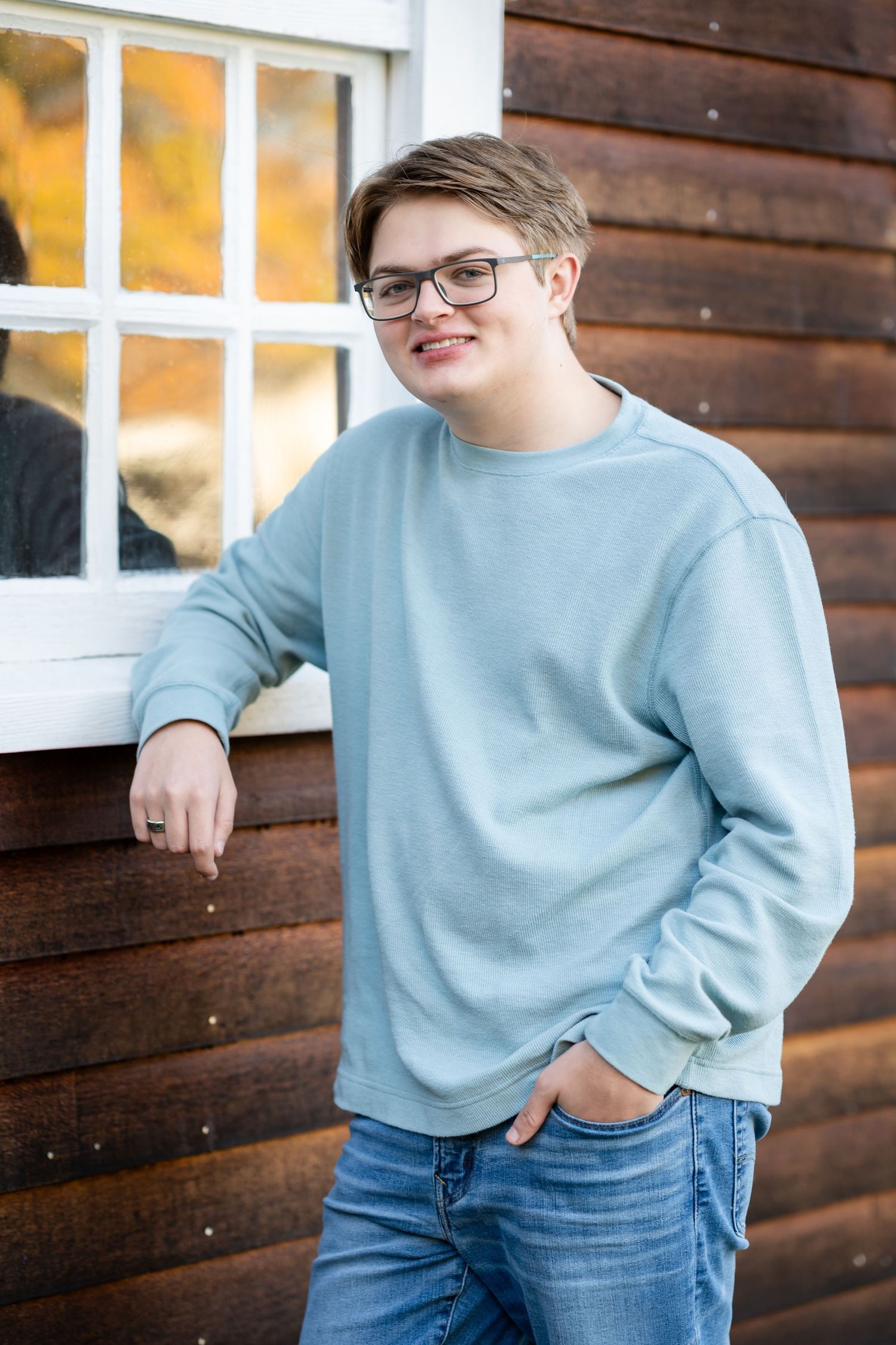 Person wearing light blue sweater and jeans poses casually against rustic wooden wall with window in background.