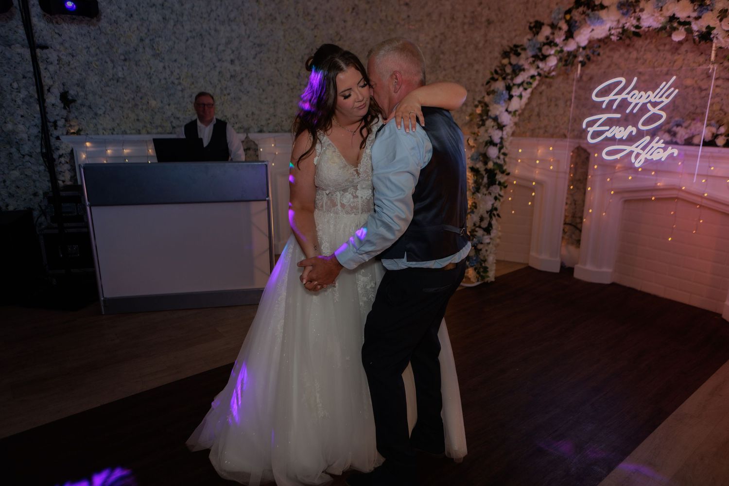 A wedding reception dance floor illuminated with pink and purple lighting under a floral arch decoration.
