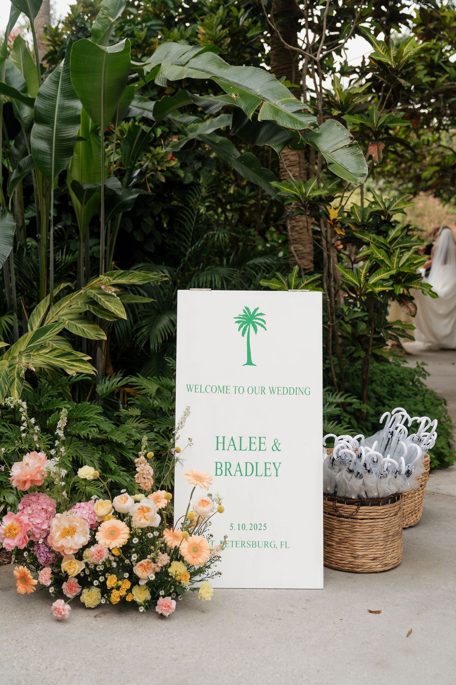 Tropical-themed wedding welcome signs with palm tree motifs and floral decorations displayed among lush greenery and roses.