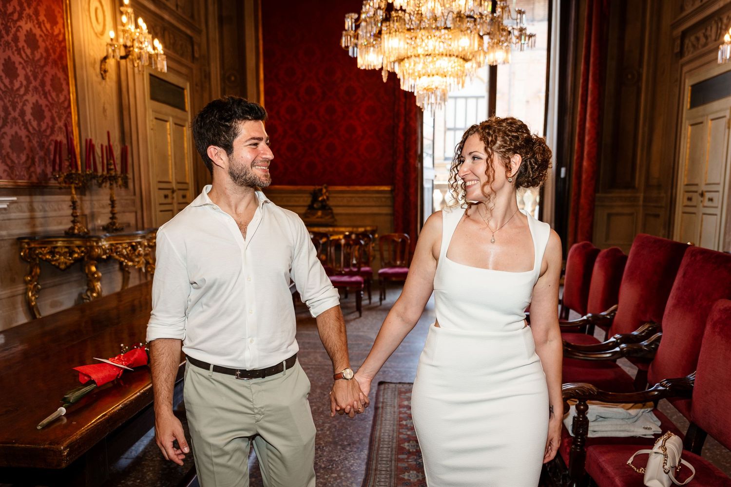 A couple shares a romantic moment in an elegant red and gold decorated room with ornate furnishings and chandeliers.