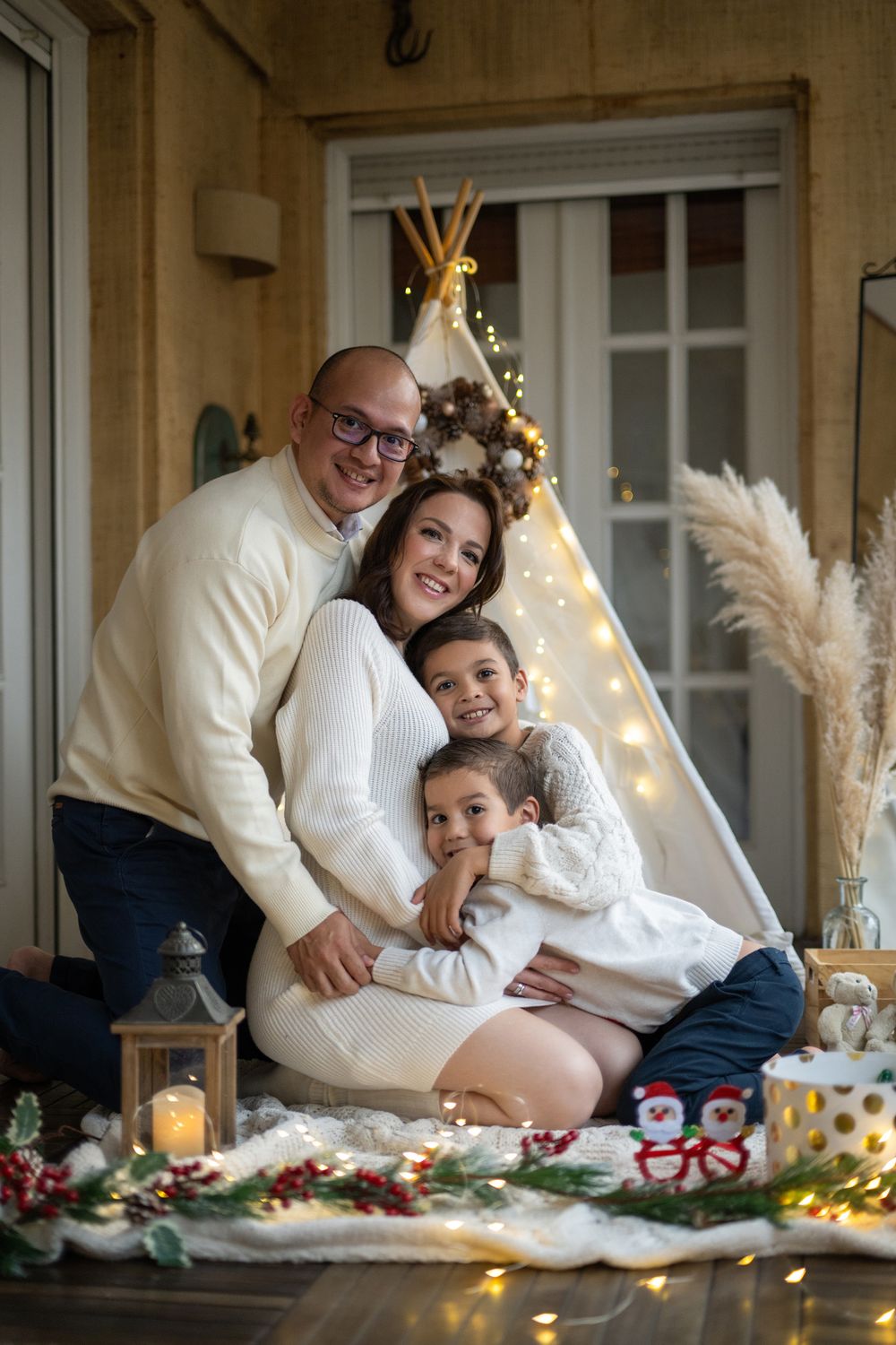 A family enjoys a cozy holiday photoshoot on the floor with twinkling lights and festive decorations in the background.