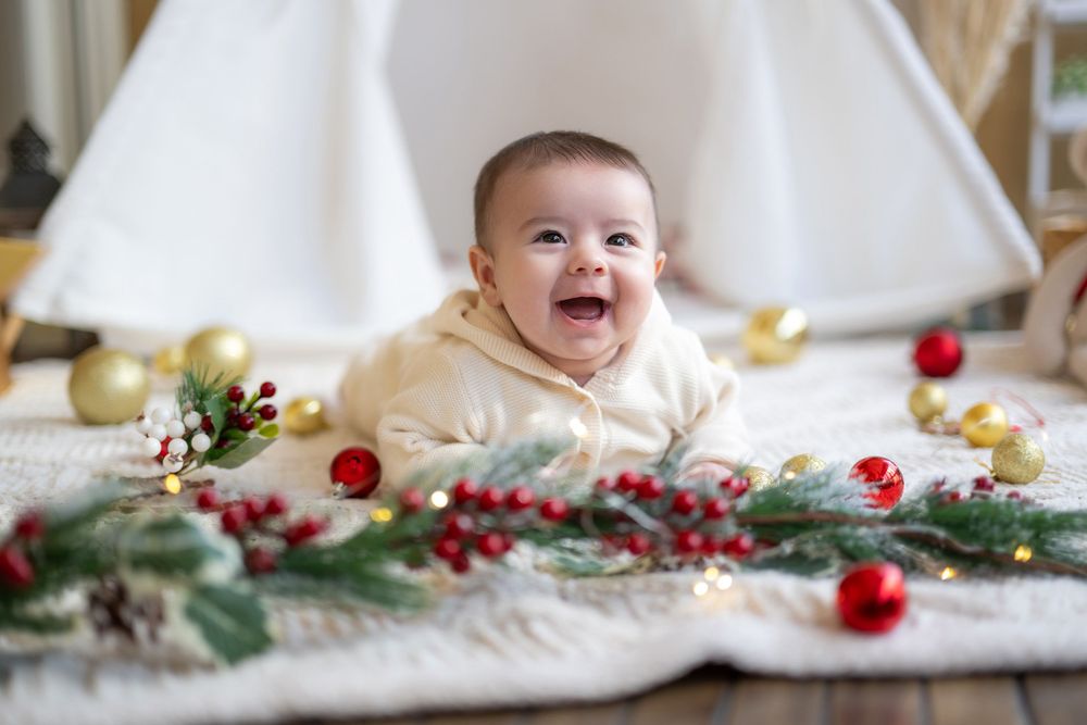A baby lies on a white blanket surrounded by festive holiday decorations including pine branches and red berries.