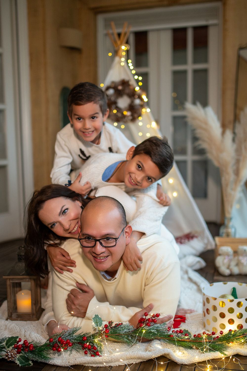 A family poses together on the floor during a cozy holiday photoshoot with festive decorations and twinkling lights.