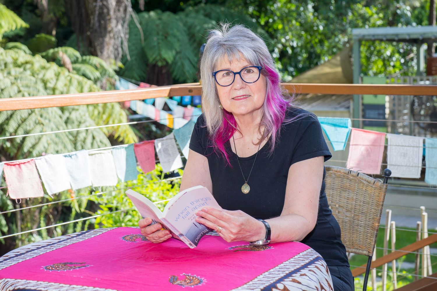 Woman with purple-tipped grey hair reads a book at a pink table in a lush garden setting.