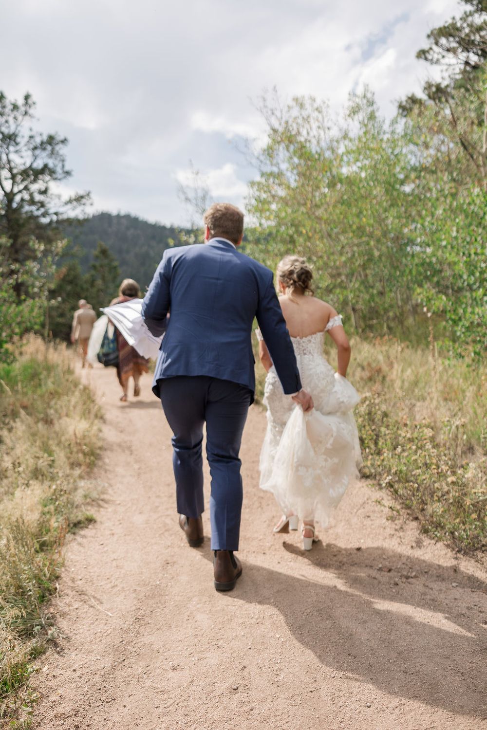 Wedding couple and guests walk along a winding mountain trail surrounded by natural scenery.